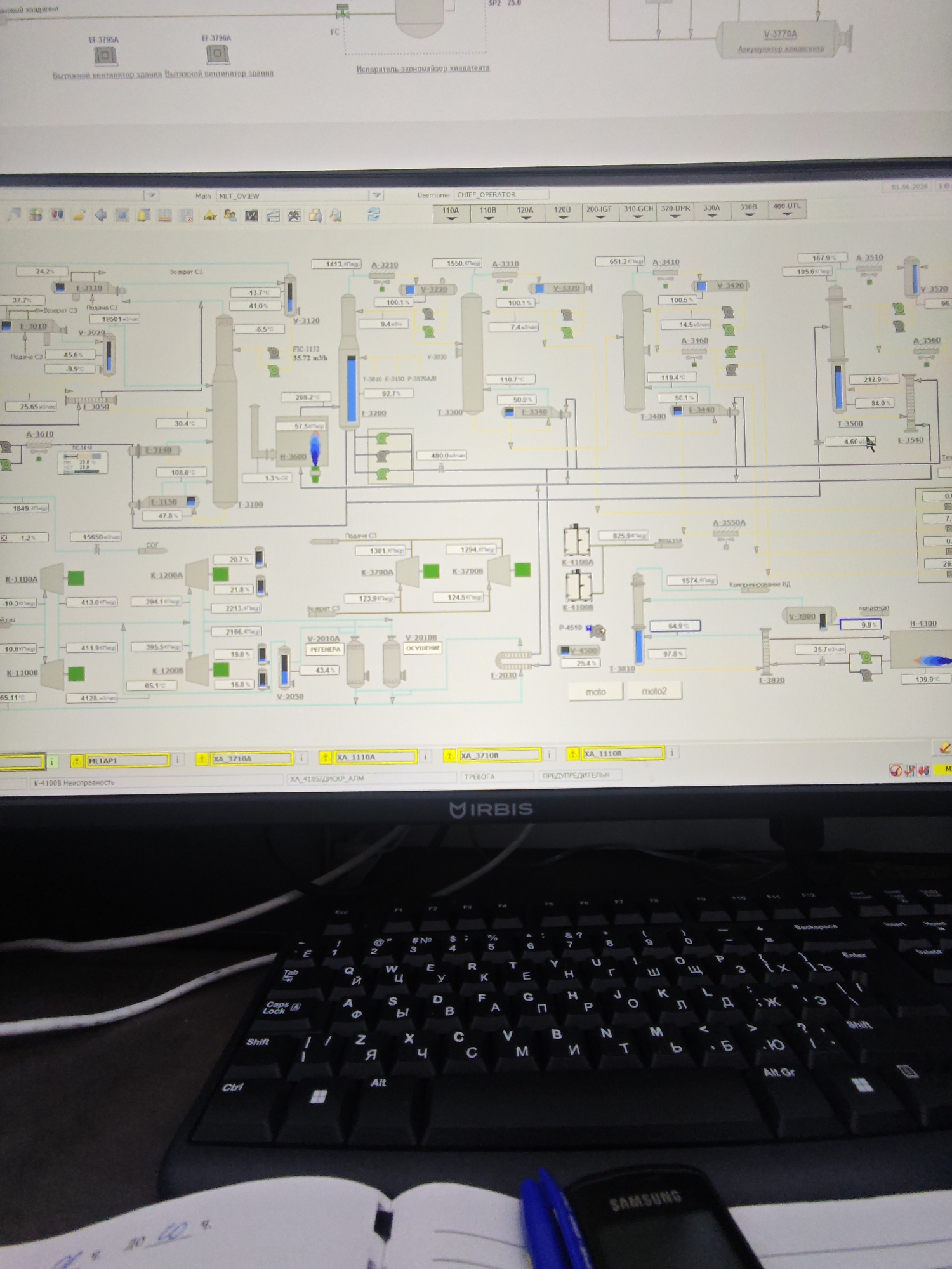 The image shows a computer screen displaying a process flow diagram, likely of a chemical or industrial plant. The diagram consists of various interconnected shapes, lines, and numerical values that represent different components and parameters of the process. The software interface appears to be in Russian.
Below the screen is a black keyboard with Cyrillic and Latin characters, indicating it's designed for both Russian and English input. A Samsung mobile phone rests on the keyboard, partially obscuring the right side. A notebook with handwritten notes is visible on the left side of the keyboard.
The environment is dimly lit, suggesting an indoor setting. The background is unclear, but appears to be part of an office or control room.
There are no people directly visible in the image. The focus is on the monitoring and control system displayed on the screen and the associated hardware.
