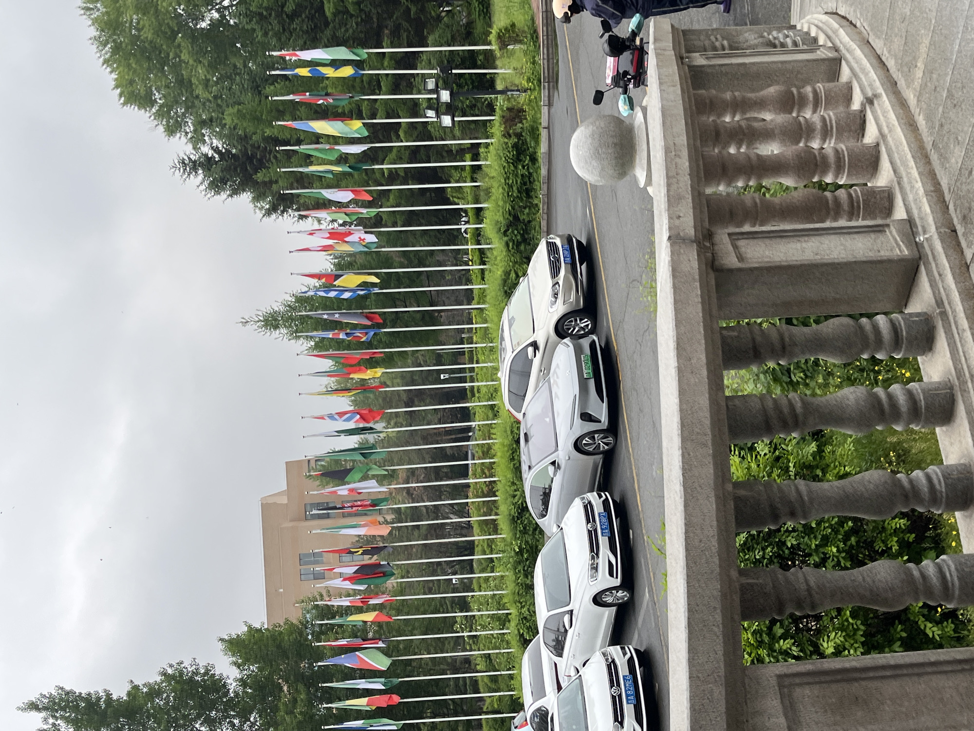 The image shows a street scene taken in Changchun City, China, from an elevated position. A row of international flags on poles lines the left side of the road. To the right of the flags, a row of white sedans is parked along the road. In the foreground, a stone balustrade indicates the vantage point, possibly a building or staircase. A person on a small scooter with a covered head is visible at the top of the image. The weather appears overcast.