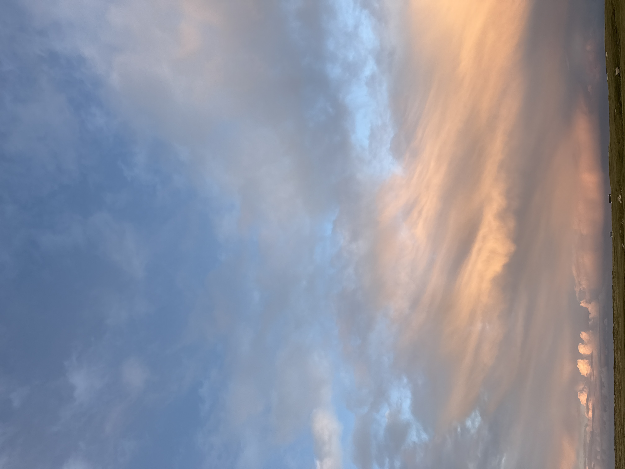 The image depicts a sky filled with clouds at an unknown location in the city of Unknown, Unknown. The left side of the sky is blue with interspersed gray clouds, while the right side shows orange-tinted clouds, likely reflecting the light of the setting or rising sun. A thin strip of green land is visible at the top right corner of the image. There are what appear to be structures barely visible on the right.