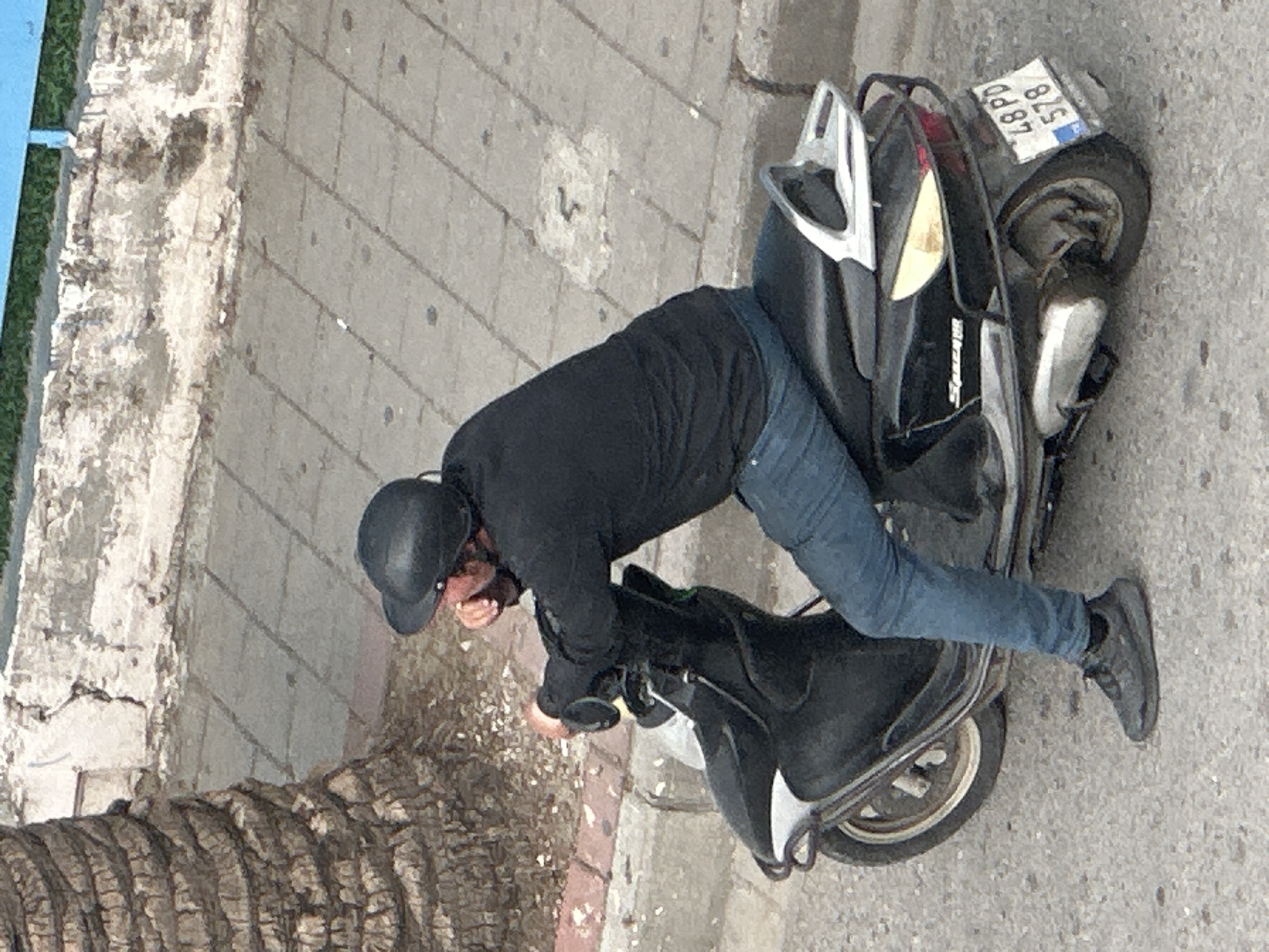 A person wearing a black helmet, a black jacket, and blue jeans is awkwardly sitting on a black scooter with a license plate reading "875 LBPD," on what appears to be a paved street. The scooter is positioned on the street next to a textured curb and a palm tree. The location is likely Fethiye, Turkey, based on the provided information. The person's posture is unusual, suggesting they are either mounting or dismounting the scooter in an unsteady manner. The surroundings appear to be a typical urban setting.