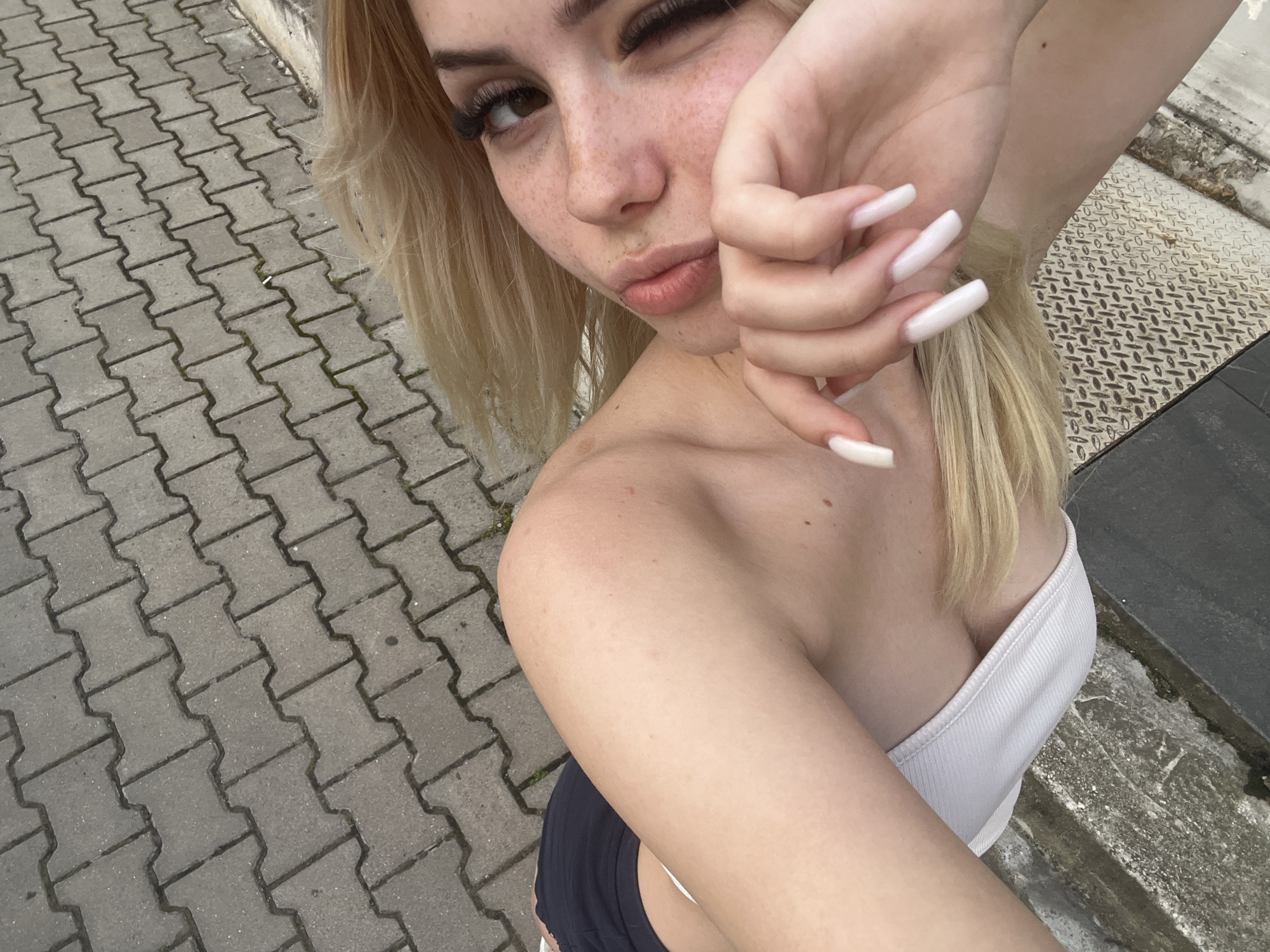 An outdoor, eye-level, medium shot shows a young woman with fair skin, blonde hair, and freckles. She is winking at the camera, her right hand resting near her face. Her lips are parted, and she is wearing a white, off-the-shoulder top. The background consists of a sidewalk made of grey interlocking pavers, a metal grate, and a grey curb. The overall setting suggests a public urban area, potentially in Plovdiv, Bulgaria.