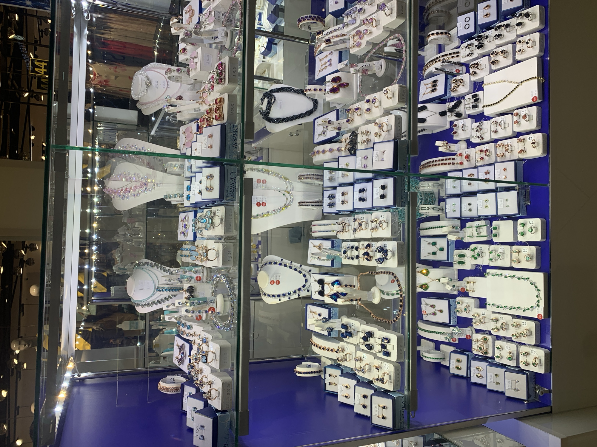 The image shows a display case filled with jewelry in a retail store. The display case is illuminated by white LED lights. On the top shelf, there are various necklaces, bracelets, and earrings, some with pink and clear stones, and others with blue stones. The middle shelf also features an array of necklaces, bracelets, and earrings, predominantly in shades of blue and silver. On the bottom shelf, the jewelry continues with more necklaces, bracelets, and earrings, including sets with green stones and other colorful gemstones. The jewelry is presented on white and blue display stands. In the background, mannequins and store fixtures are visible. The overall impression is of a well-organized jewelry store display.