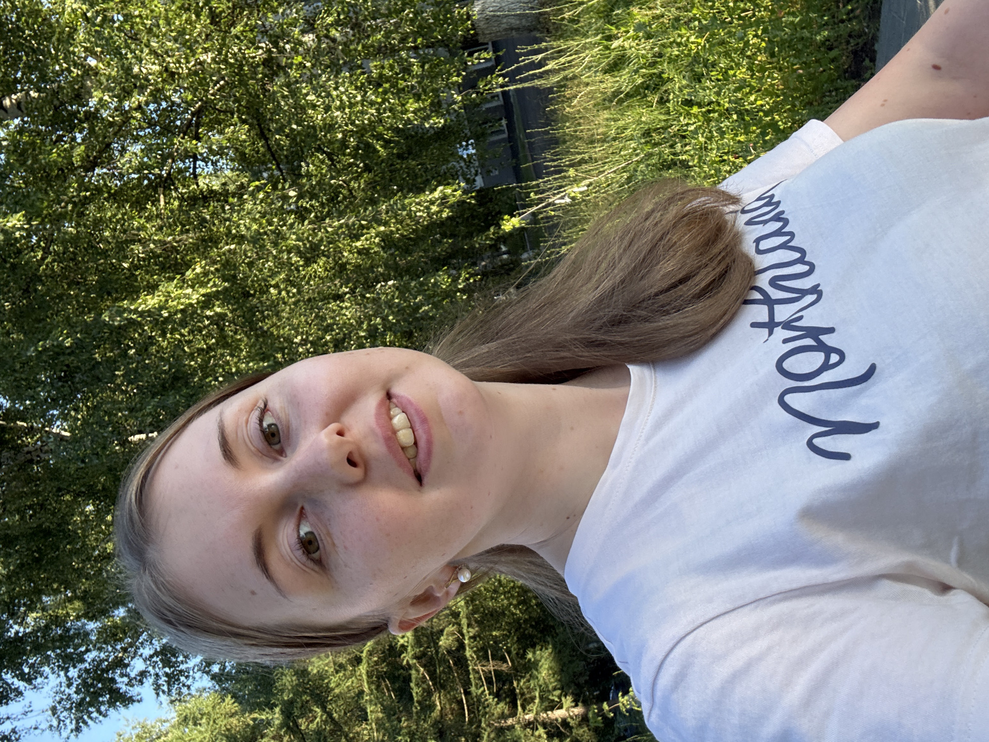 In an outdoor, medium-shot, selfie-style photograph, a young woman with light brown hair, freckles, and green eyes smiles and looks at the camera. She is wearing a white t-shirt with dark blue lettering that reads "Northland." She has a small pearl earring in her left ear. The background consists of green trees and tall grass, with a building visible in the distance. The scene is illuminated by natural sunlight.