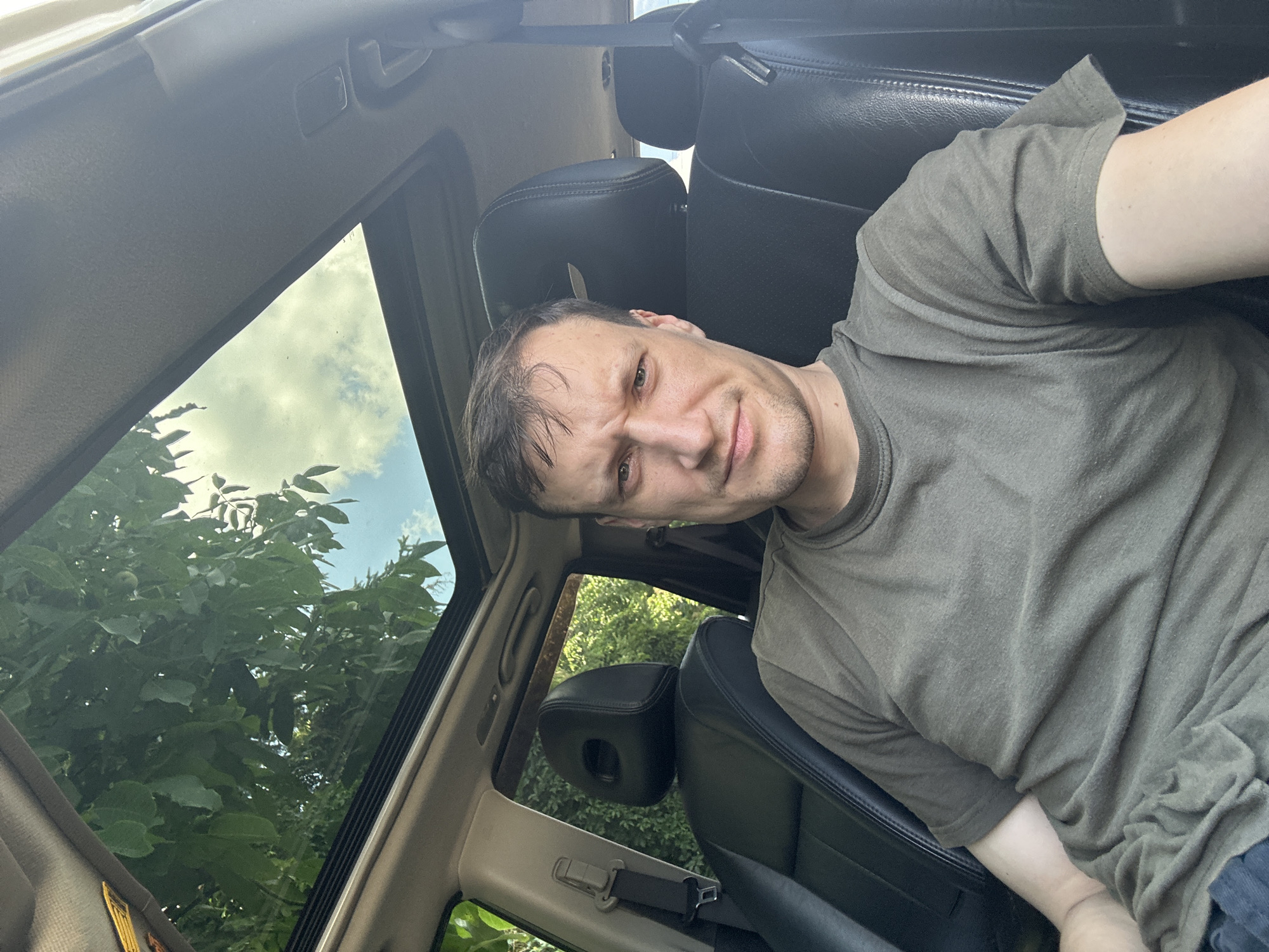A man with short brown hair and a strained expression is taking a selfie inside a car with a sunroof open, revealing green trees and a blue sky with clouds. He is wearing a dark green t-shirt and is seated in the driver's seat. The interior of the car appears to be black leather. The location is the city of Shatava, Ukraine.
