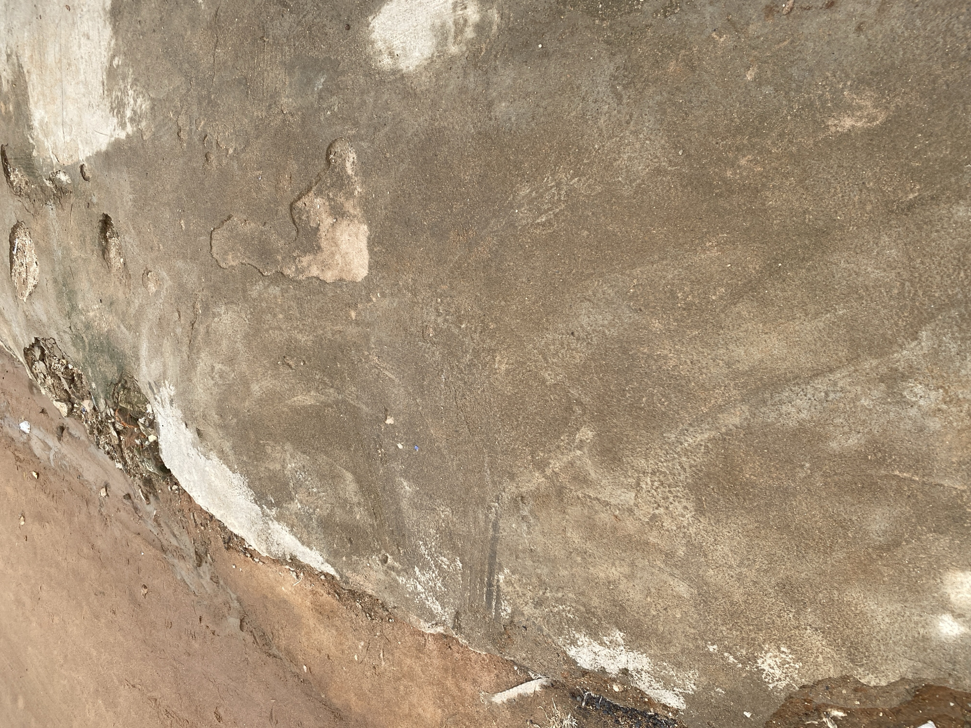 This is a close-up, top-down view of a textured, weathered concrete wall with adjacent sandy ground. The concrete surface is predominantly a mottled brown and gray, showing signs of age and wear. There are patches of lighter, almost white material, possibly a faded paint or plaster, and darker, damp-looking streaks. The texture is uneven, with subtle undulations and small pits.
On the left side of the image, the concrete wall meets the ground, which is a dry, reddish-brown sandy material. This transition zone reveals the rough, uneven base of the wall, with some loose stones and debris visible. The sandy ground appears somewhat disturbed, with a few faint imprints or marks.
The lighting suggests it is daytime, with the sun casting some subtle shadows and highlighting the textures of the concrete and sand. There are no visible people or specific objects that would indicate an activity or event. The scene appears to be an outdoor location, possibly an exterior wall or a section of a construction site. The overall impression is one of neglect and the passage of time, with no discernible emotions conveyed. No text is visible in the image. The context provided indicates this scene is located in Jalingo, Nigeria.