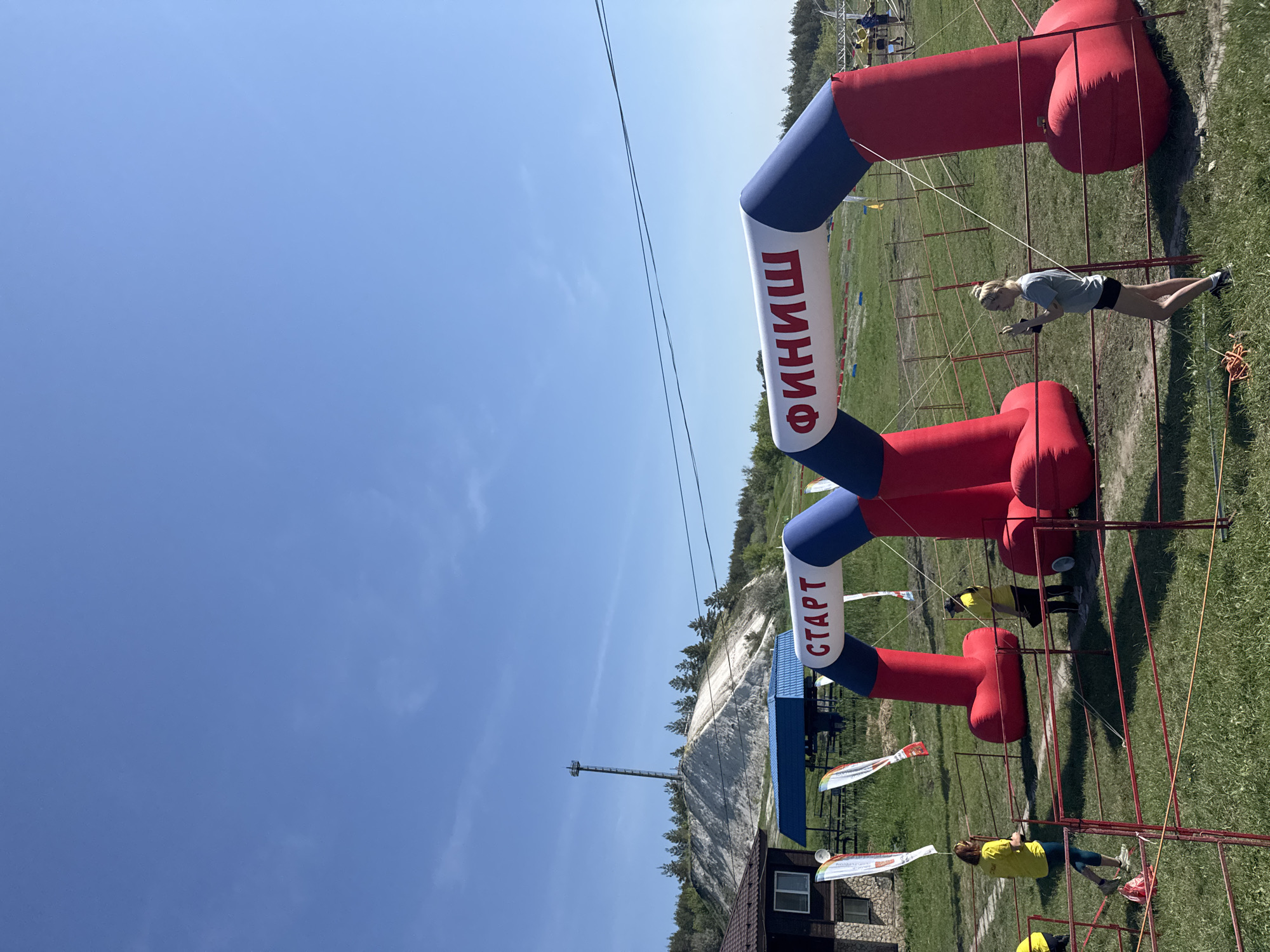 The image depicts an outdoor scene in Khvalynsk, Russia, showing the start and finish line inflatables for an event, with people interacting with them. The sky is clear blue. The inflatables are red, white, and blue and have the words "СТАРТ" (start) and "ФИНИШ" (finish) written on them in Russian. People are either assembling or disassembling the inflatable archways. To the left, there is a building with a blue roof, and in the background, there is a forested area and a ski jump ramp. There are other people scattered around the grassy area.