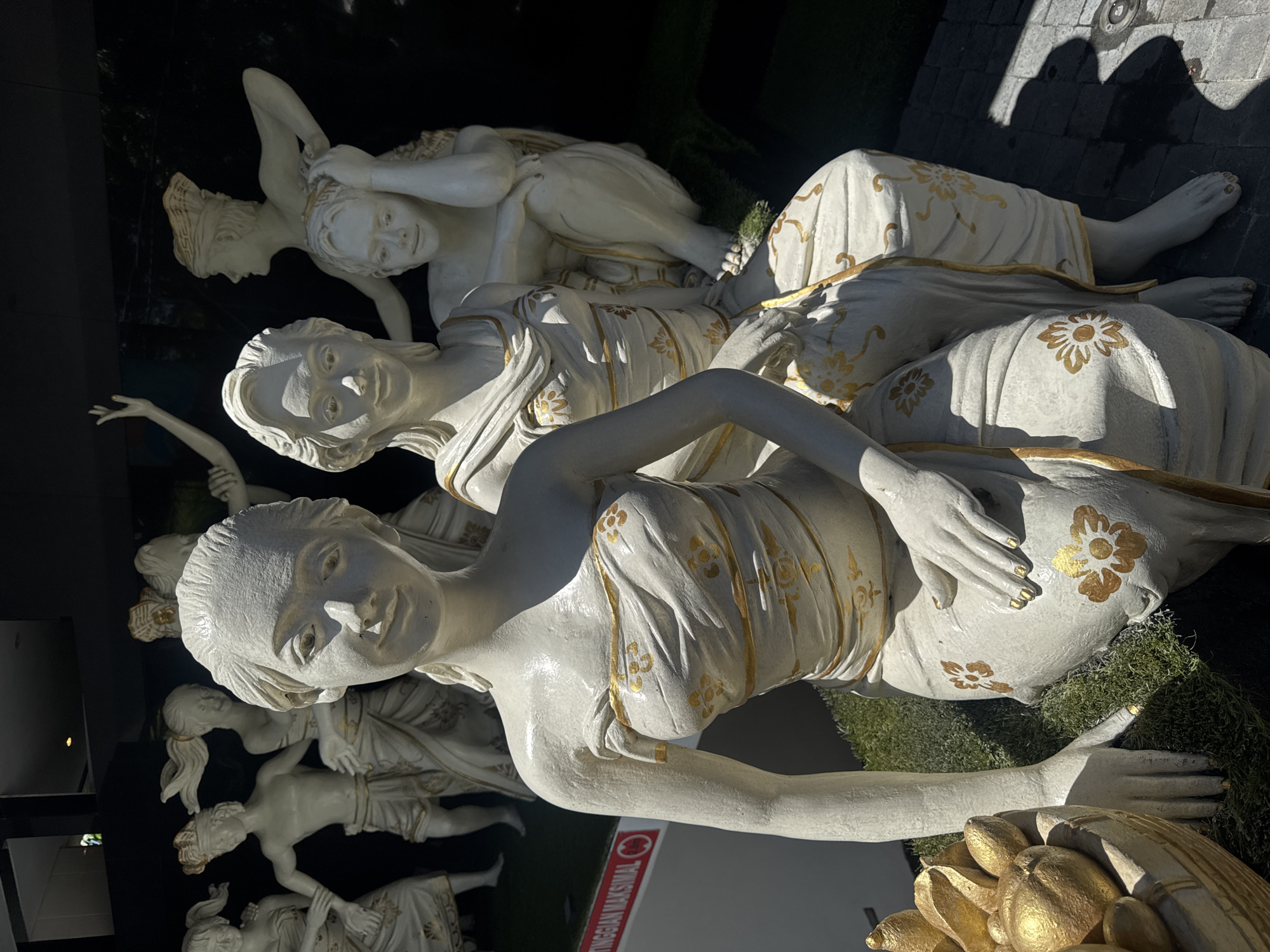 The image depicts a group of white statues of reclining women adorned with gold accents, displayed on a dark surface with patches of green moss. The scene is partially shadowed, with the background showing a glimpse of dark structures and possible vegetation. The statues are arranged in layers, with figures both reclining and partially raised. Text is visible, reading "TINGGIAN MAKSIMAL." The scene is located in Kuta, Indonesia.