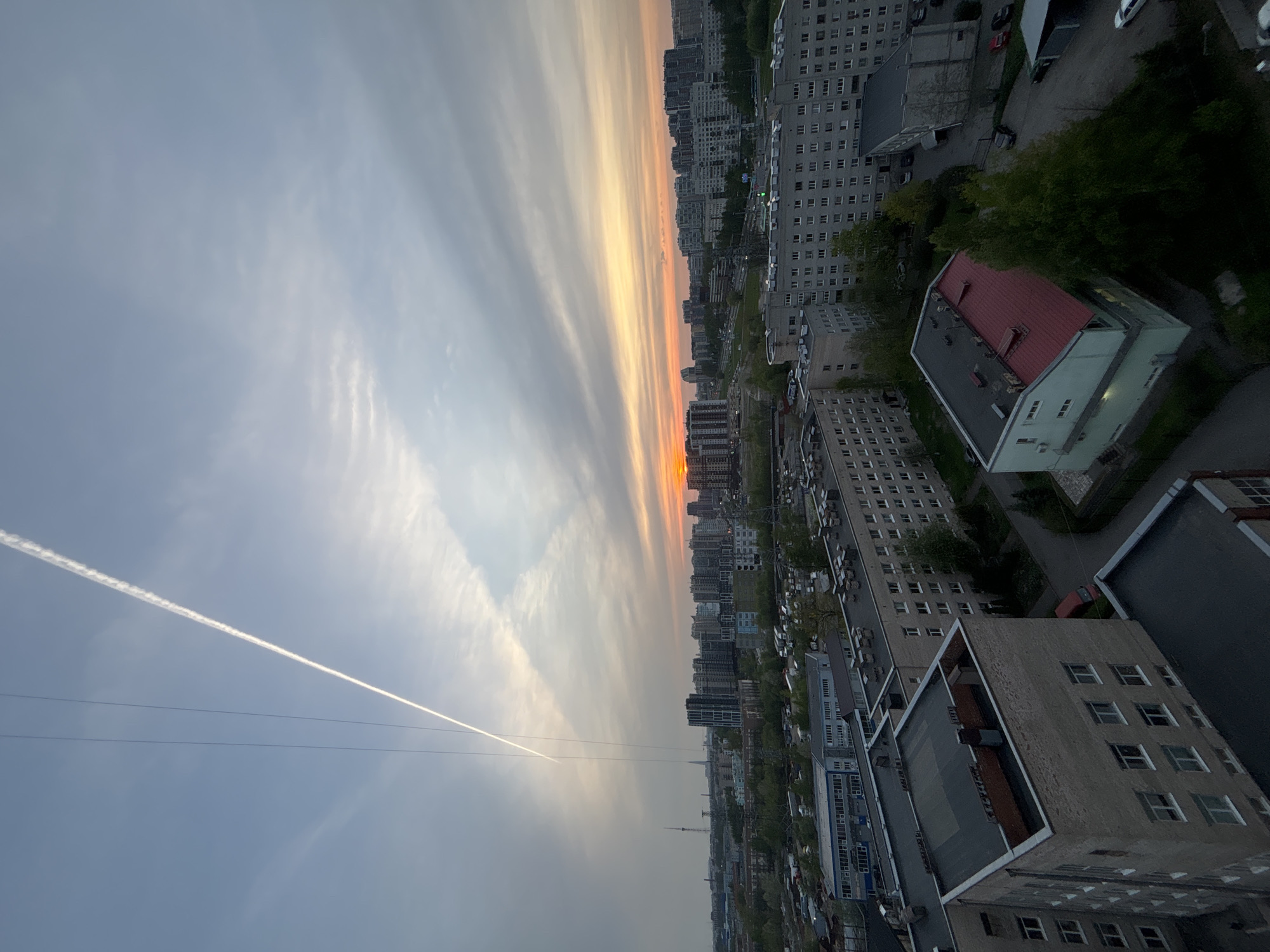 The image shows a cityscape at sunset. The view is from a high vantage point, possibly a building. The sky is split with shades of blue and orange indicating the time of day is sunset. There is a contrail bisecting the sky from lower left to near the center.
Below the sky is a dense urban area comprised of mid-rise buildings. A street runs parallel to the line of sunset. A building with a red roof can be seen in the foreground. The presence of cars parked along the street suggests activity. The environment appears urban with a mix of buildings and a few trees.