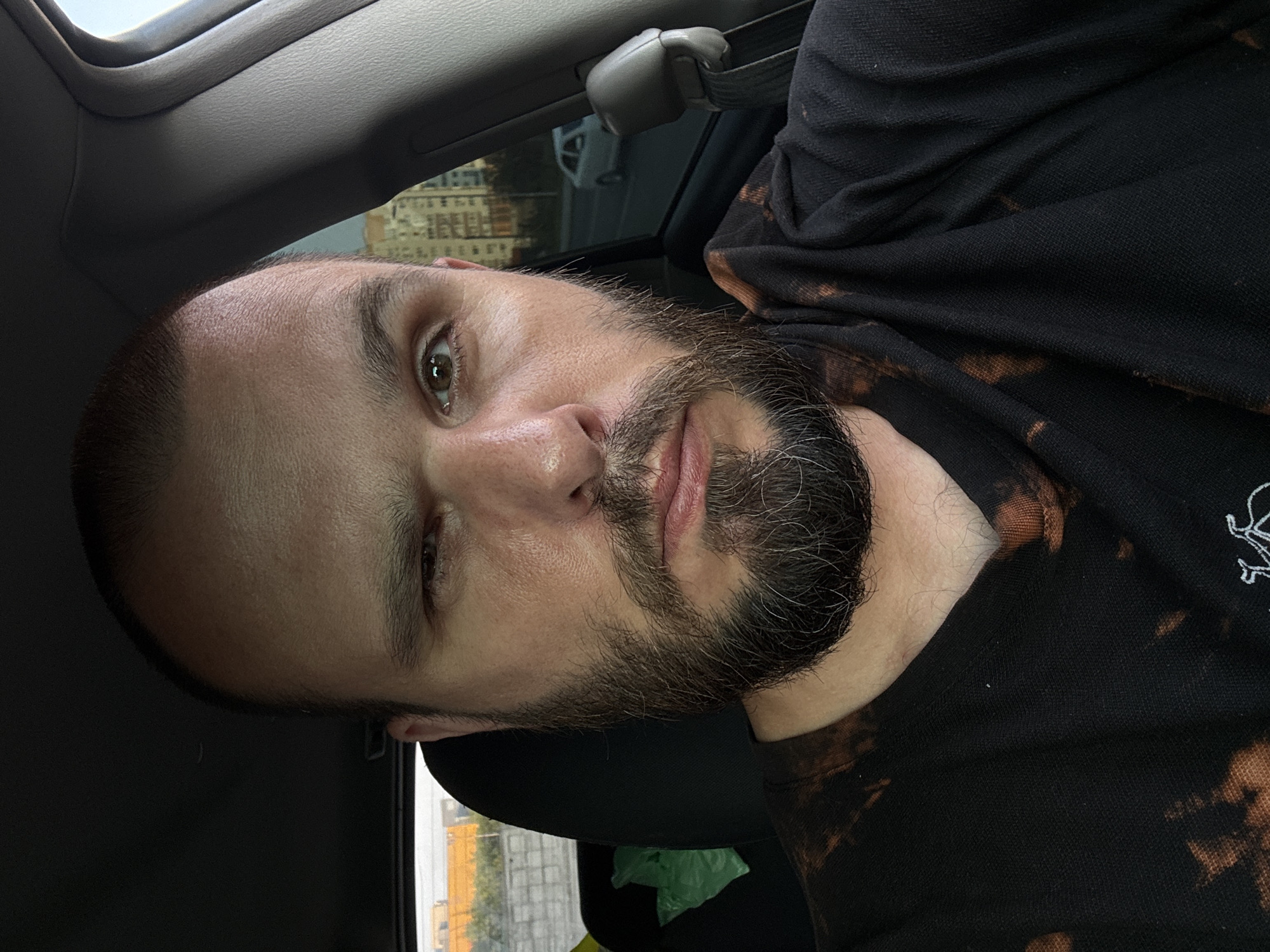 A medium shot portrait of a man in his 30s, seated in the driver's seat of a car. The man has a shaved head, brown eyes, and a full beard. His right eye is wide open, while his left eye is partially closed in a wink. He is wearing a black t-shirt with a tie-dye bleach pattern. The car interior is visible, including a dark headliner and a grey plastic trim piece on the right side. Outside the car window, a street with buildings and a white car can be seen. The scene appears to be in an urban environment, likely Nizhny Novgorod, Russia, given the architectural style of the buildings. The lighting suggests it is daytime.
