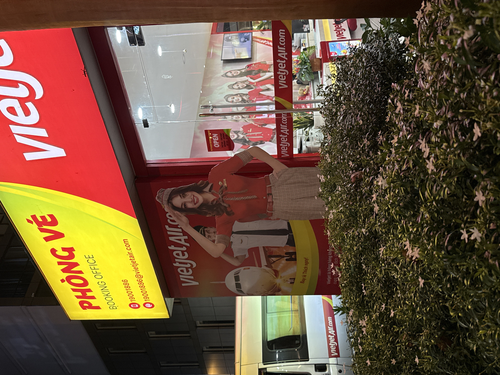 A VietJet Air office in Thủ Đức, Vietnam is partially obscured by a bush. The sign states "Booking Office" in English and Vietnamese. The VietJet Air advertising contains a female model in an air-stewardess uniform and the phrase "Bay là Thích ngay!". A television screen in the office displays images of the flight attendants. A service van is visible. The office sign is yellow and red. The address and phone numbers are printed in red.