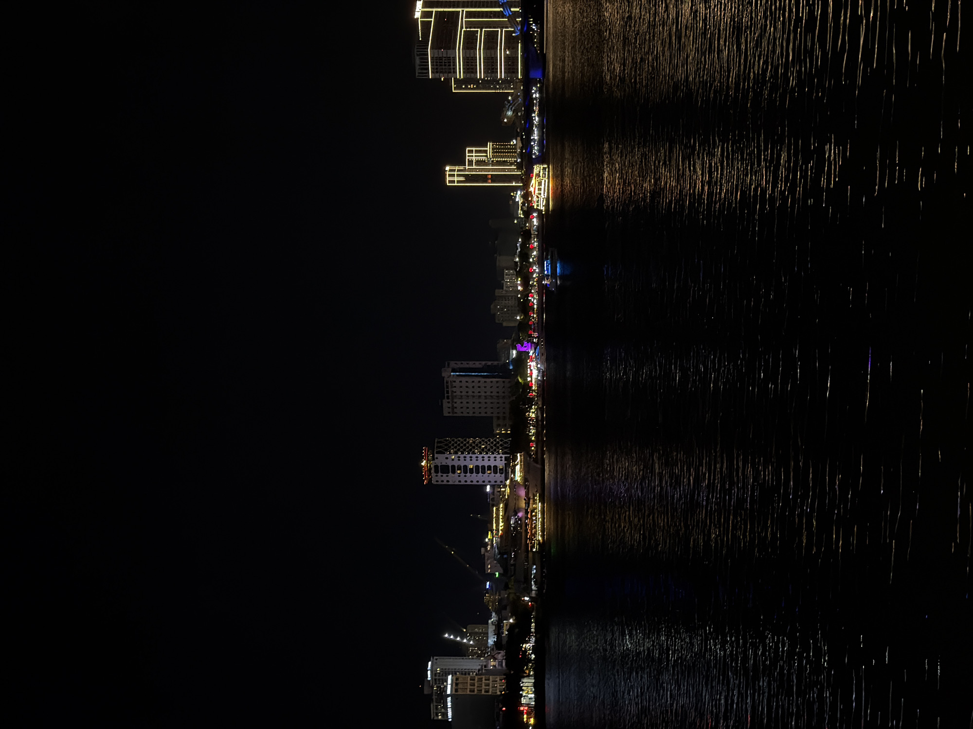 This is a nighttime, vertical shot of the Đà Nẵng coastline. The image depicts a cityscape with numerous illuminated buildings, including hotels and high-rises, lining the waterfront. The lights from the buildings cast reflections on the dark water, creating a shimmering effect. Several boats are visible on the water, some also illuminated. The overall scene suggests a vibrant and active urban environment at night. The time of day is clearly night, and there are no visible weather cues. The location is identified as Đà Nẵng, Vietnam. There is no discernible text in the image. The atmosphere appears lively and modern due to the extensive lighting.