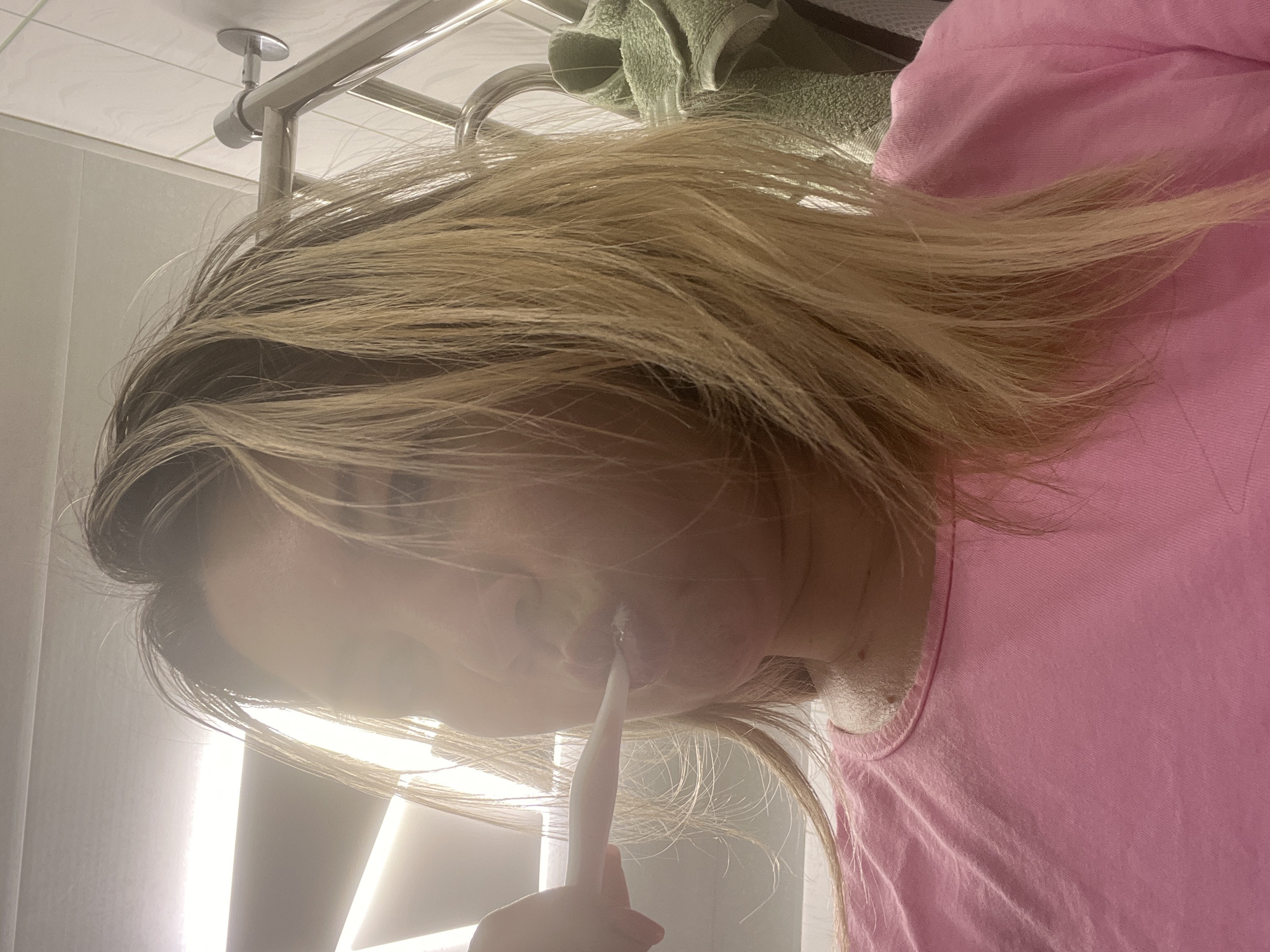 A woman with blonde hair is brushing her teeth in a bathroom. She is wearing a pink t-shirt. A towel is hanging on a rack in the background. The scene appears to be in Sochi, Russia.