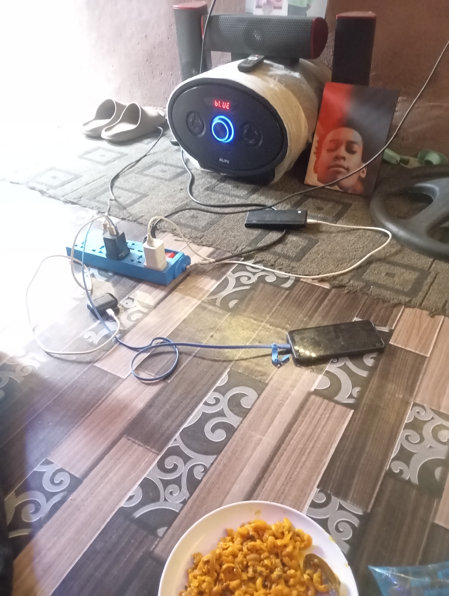 This is an indoor, eye-level photograph depicting a cluttered yet functional floor space. The setting appears to be a casual room, possibly a living area or bedroom, characterized by a distinct floor pattern combining wood-like planks and decorative grey and white swirling motifs. A patterned rug or carpet is visible in the background, underneath several electronic devices.
In the center of the image, a black and blue portable speaker is prominently displayed, its circular central light glowing blue and a small screen displaying the text "BLUE." The brand name "ALIPU" is also visible below the screen. A small remote control rests on top of this speaker. To its left, a pair of light-colored slide sandals is placed on the rug. To the right of the speaker, a printed photo of a young male with a red and black background leans against an unseen object. Further back, two smaller black and red speakers are partially visible.
A blue power strip is situated on the floor, connected to various chargers and cables. A white cable extends from the power strip to a black rectangular device, possibly a USB hub or external hard drive. Another blue cable connects this black device to a black smartphone, which is lying flat on the floor and appears to be charging. Other cables are tangled around the power strip.
In the foreground, a white bowl filled with a yellow/orange pasta dish, likely macaroni with sauce, sits on the floor. A spoon is visible in the bowl. A clear or light blue plastic bag is also partially visible near the food. A black weight plate is partially seen on the right side of the image, resting on the rug.
The room is brightly lit, with a strong light source coming from the left side of the frame, creating some glare and highlights. The overall scene suggests an informal moment of relaxation or daily activity, with food and entertainment easily accessible.