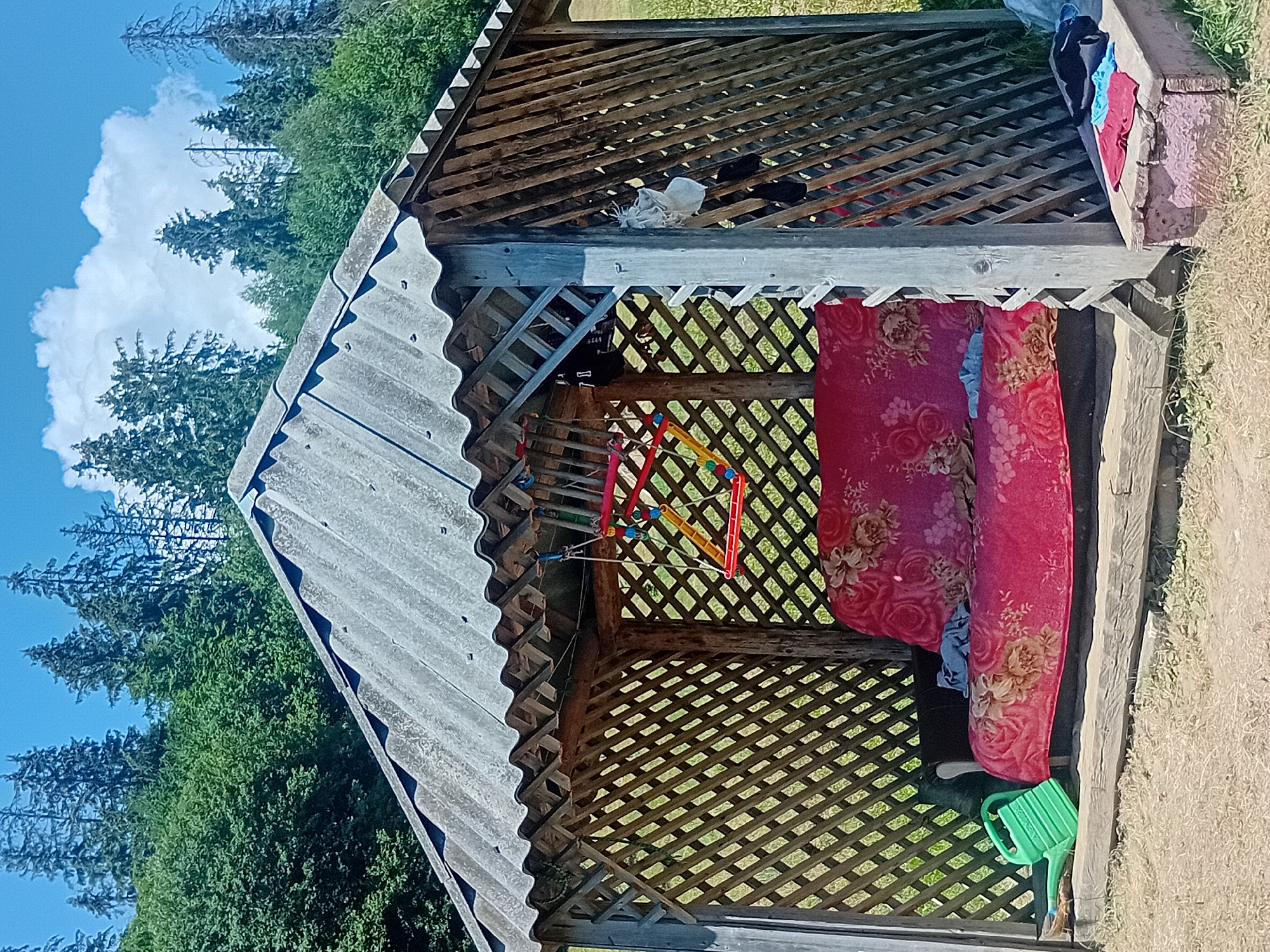 In Verkhniachka, Ukraine, a weathered wooden gazebo stands under a bright, partly cloudy sky. The gazebo's roof is made of corrugated asbestos cement sheets. Inside, a red floral-patterned couch or futon is covered with a patterned throw blanket. A colorful, abstract-shaped toy is suspended by strings from the gazebo's ceiling. To the left of the couch, a bright green plastic watering can rests on the ground. The gazebo's walls are constructed with diamond-shaped lattice wood panels. Outside the gazebo, on a wooden crate to the right, a pile of clothes, including a red, blue, and black garment, is visible. The surrounding environment appears to be a rural or forested area, with green trees and a grassy ground.