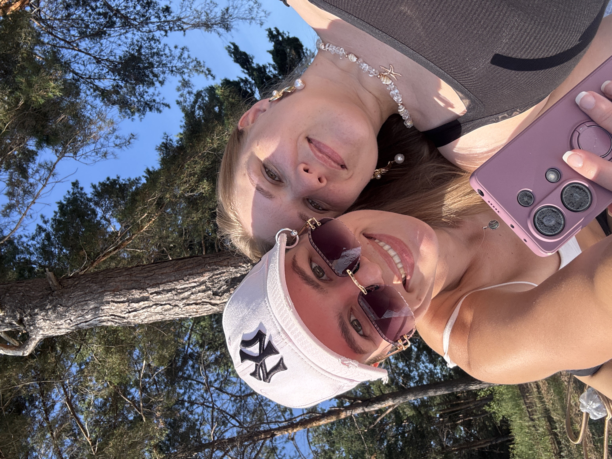 This is a selfie of two young women outdoors. The woman in the foreground is wearing a white baseball cap with a black "NY" logo, sunglasses, and a white bikini top. She is smiling and holding a light purple phone. The second woman is in the background, wearing a dark tank top and a beaded necklace with a starfish charm. She is also smiling. The image is taken from a low angle, looking up towards the sky, with trees and blue sky visible. The lighting suggests it is daytime, likely sunny. The overall mood appears cheerful and relaxed.