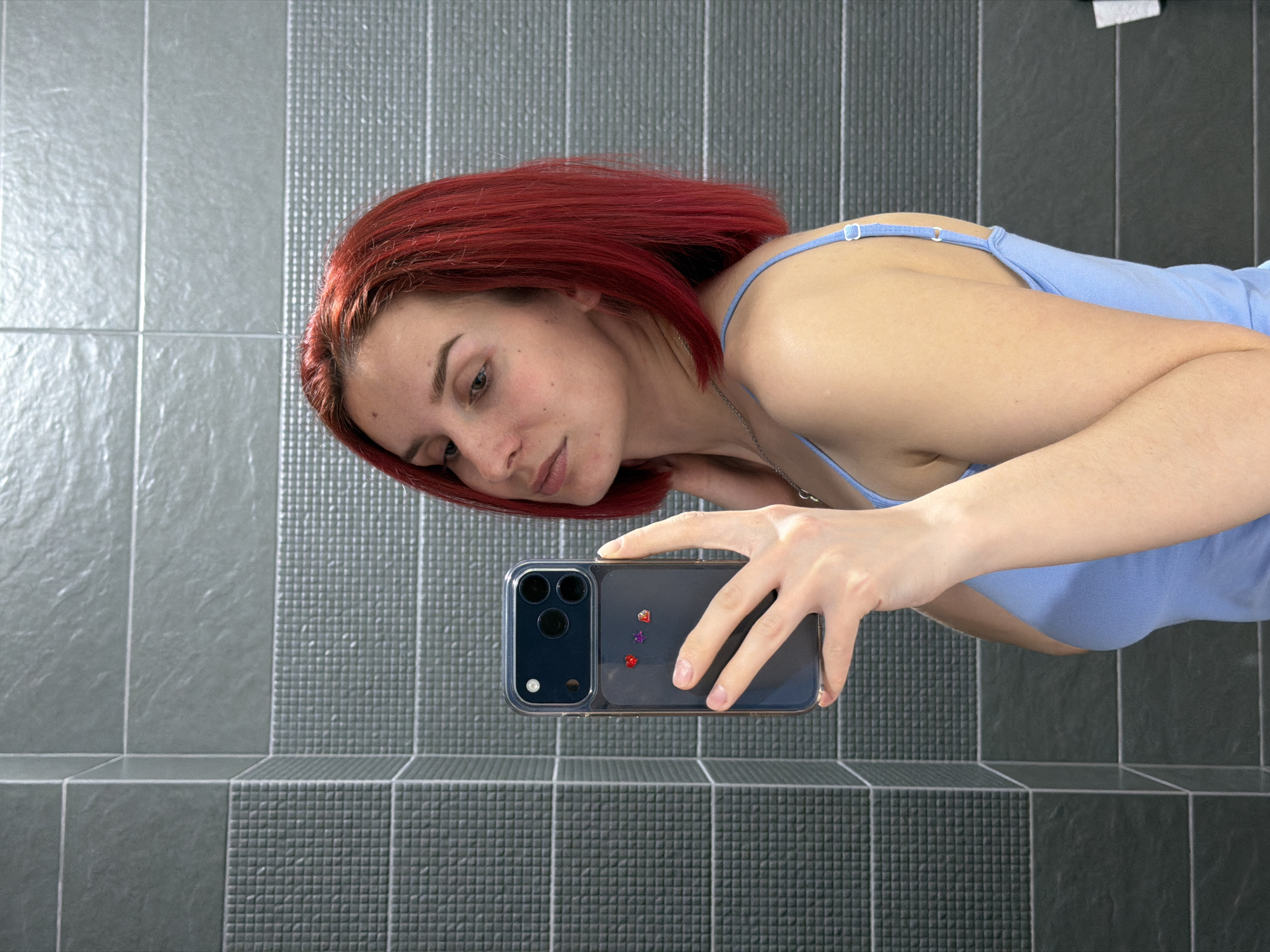 A woman with red hair and a blue tank top is holding a cell phone in her right hand, apparently taking a selfie. The phone is an iPhone with a clear case decorated with small, colorful stickers. The background is a tiled wall, possibly in a bathroom. The location is presumed to be Chaikovsky, Russia, based on the user prompt.