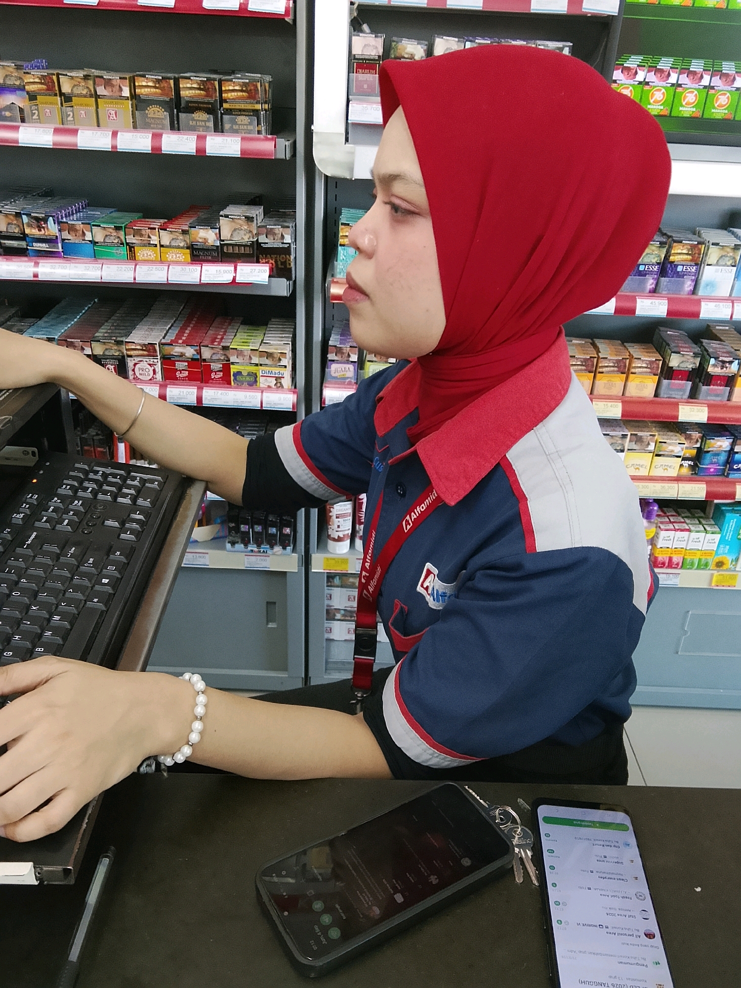 In the city of Talise, Indonesia, a person wearing a red hijab and an Alfamidi uniform is working at a counter, using a keyboard. The person's left hand, adorned with a pearl bracelet, is positioned over the keyboard. To the right of the keyboard, a smartphone rests on the counter. The background reveals shelves stocked with various products, including cigarettes, typical of a convenience store. The lighting suggests an indoor environment, likely during operating hours.