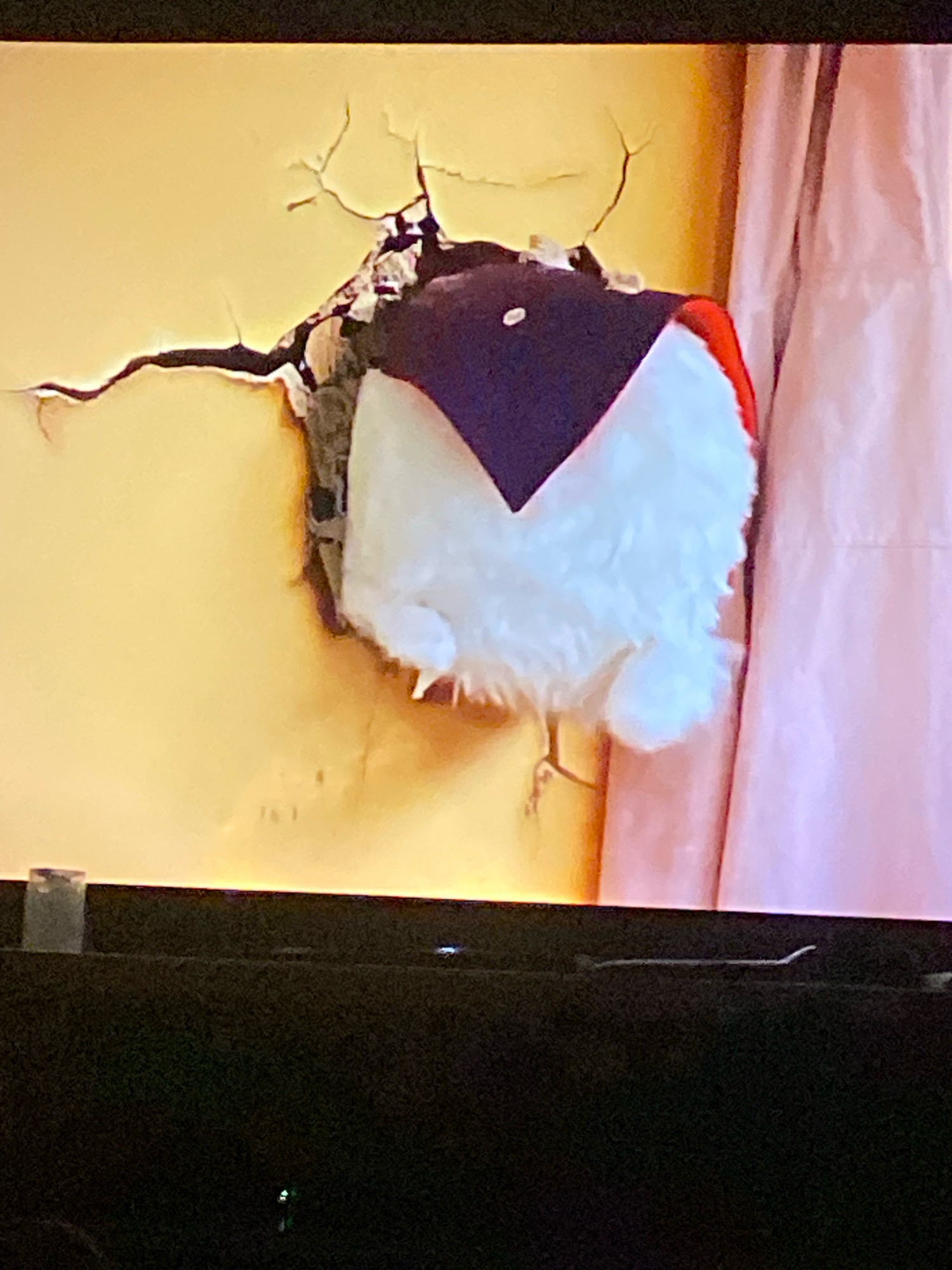 The image shows a stuffed toy, possibly a penguin, that has broken through a cracked yellow wall. The toy is mostly white with a dark blue triangular section on its head and a red accent on its head. The wall appears to be textured and has significant cracks radiating from the hole. To the right of the wall is a pale pink curtain. The lower portion of the image shows the edge of a black screen, likely a television, with some subtle reflections. There is no discernible location in the city of Shahtinsk, Kazakhstan provided in the image.