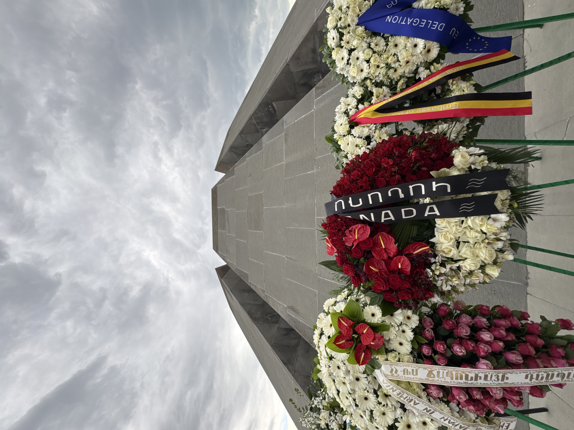 The image shows a gray concrete structure, possibly a monument, adorned with multiple floral wreaths. The wreaths are composed of various flowers including white daisies, red anthuriums, and pink roses. Ribbons with lettering in different languages are attached to the wreaths. One ribbon reads "EU Delegation" and another has the flag of Belgium and the words "Embassy of the Kingdom of Belgium." A black ribbon has the word "Canada" in English and Armenian. The monument is likely located in Yerevan, Armenia, considering the lettering on one of the ribbons includes Armenian. The background features a cloudy sky.
