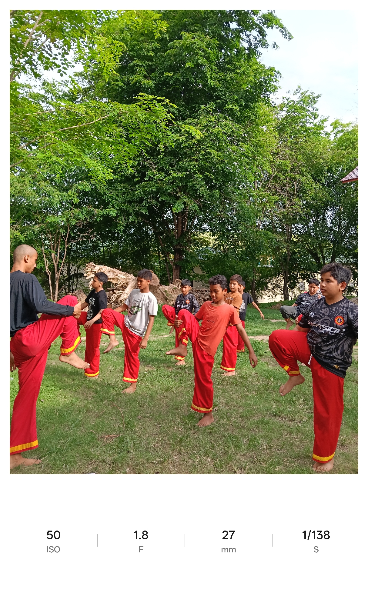 The photograph captures a martial arts training session taking place outdoors, presumably in Lhokseumawe, Indonesia. A group of approximately 8-10 individuals, mostly young males, are dressed in red pants with yellow trim, a traditional uniform style. They are barefoot and appear to be practicing a synchronized kicking motion. One of the individuals, possibly an instructor, stands at the front of the group wearing a black shirt. The setting is a grassy area under the shade of large trees. Behind the practitioners, there's a pile of what looks like cut tree branches or logs. The image's metadata indicates an ISO of 50, an aperture of f/1.8, a focal length of 27mm, and a shutter speed of 1/138 second.
