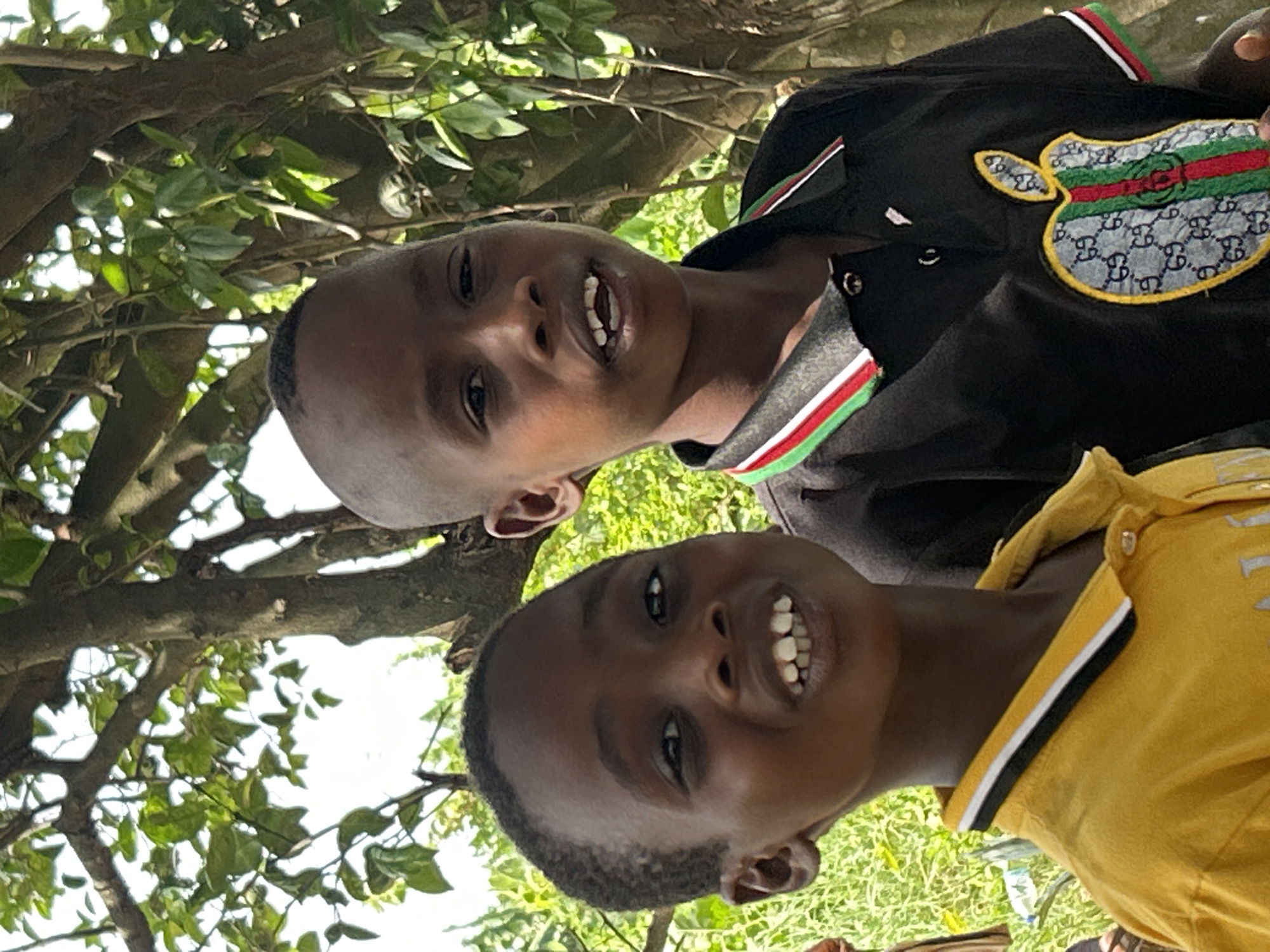 Two boys are visible in the image, possibly standing outdoors under a tree. The upper boy is wearing a black shirt with a colorful apple-shaped patch. The lower boy is wearing a yellow shirt with white accents. The location is not discernible from the image.