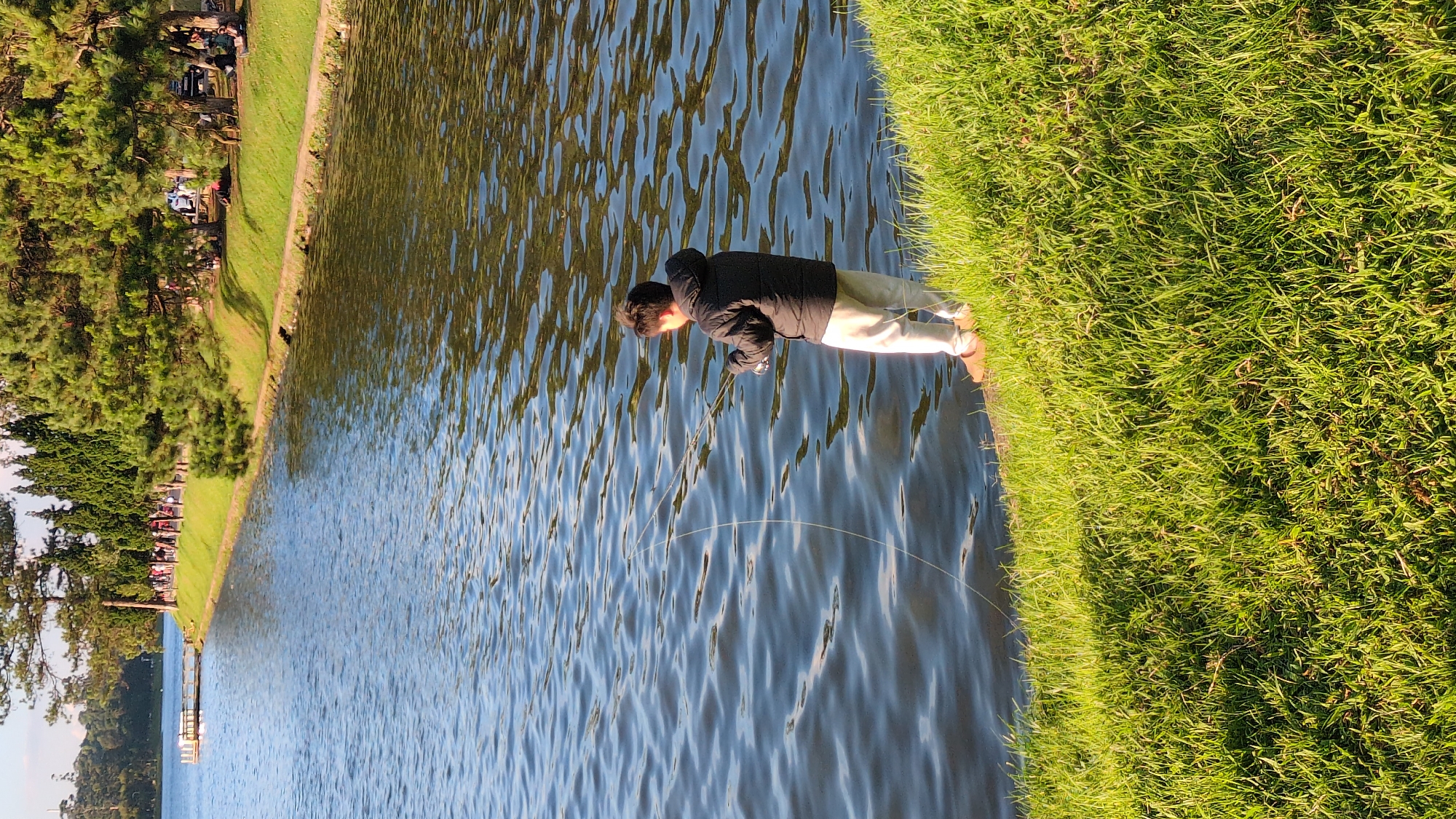 A young boy stands on the grassy bank of a lake in Da Lat, Vietnam, holding a fishing rod and fishing. The boy is wearing a dark puffer jacket, light-colored pants, and brown shoes. The lake is calm with gentle ripples, reflecting the sunlight. In the background, a paved path runs along the lake, bordered by lush green grass and pine trees. Several people are seen walking along the path and sitting on the grass under the trees, enjoying the scenery. A small wooden dock extends into the lake on the left side of the image. The overall scene depicts a peaceful outdoor setting.