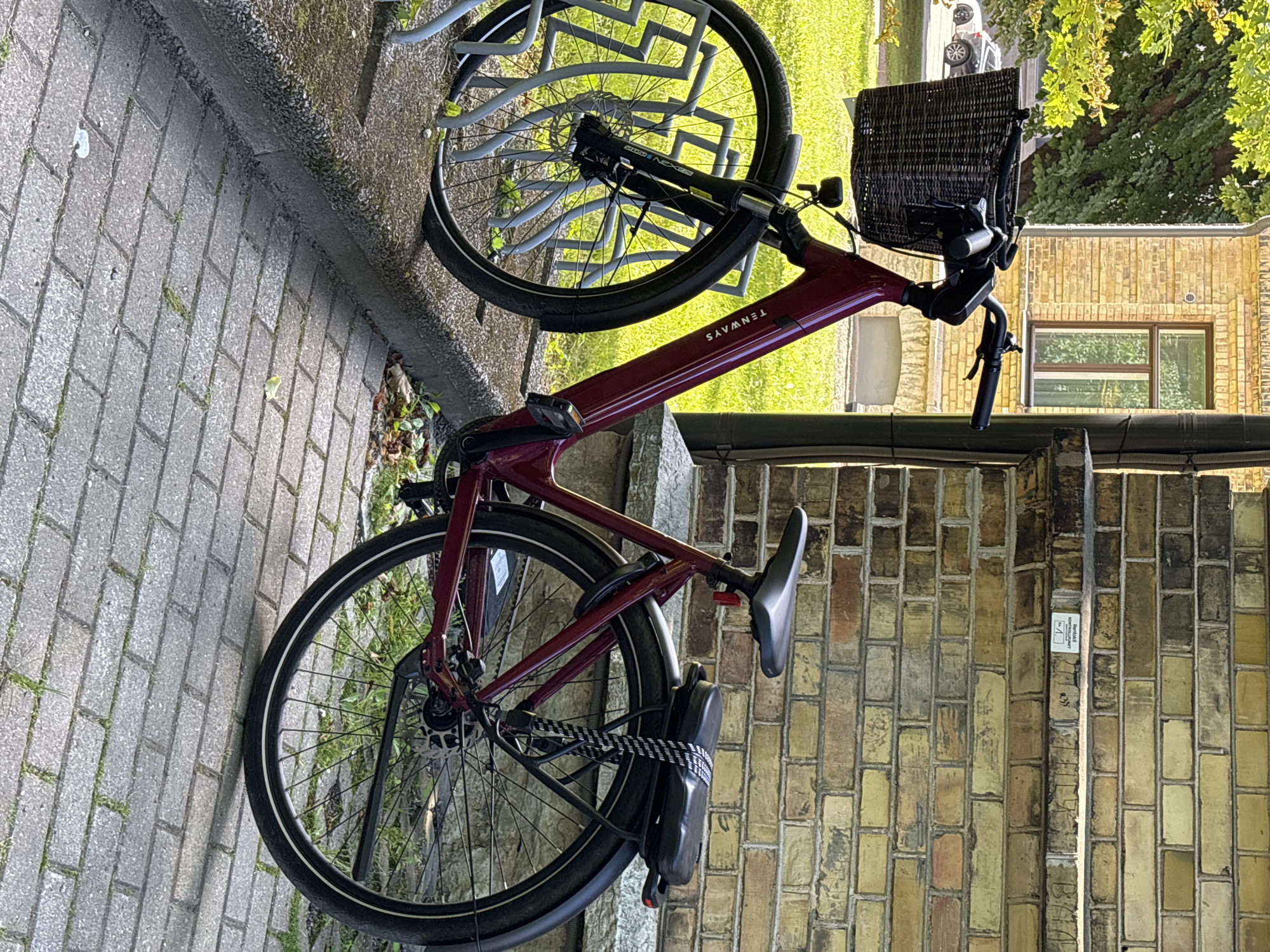 A maroon Tenways bicycle is parked in a bike rack in front of a yellow brick building in Tallinn, Estonia. The bicycle has a wicker basket on the handlebars and a black seat on the rear rack. A black and white striped lock is attached to the rear rack. The ground is paved with gray bricks. The building is visible in the background with a green tree.