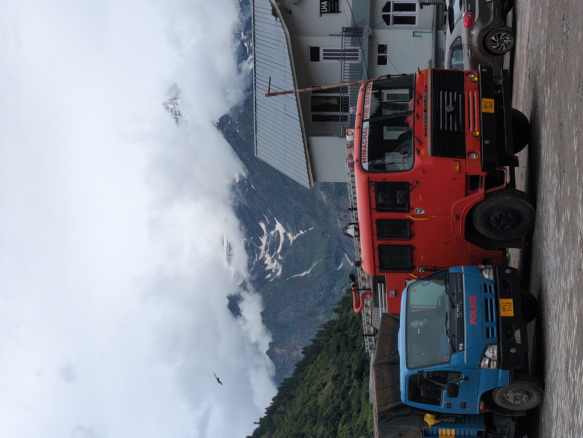 In Keylang, India, on a cloudy day, a blue police truck and a red fire truck are parked next to a building. The building has a gray corrugated roof and white walls with balconies and windows. Mountains, some with snow, are visible in the background. A bird is flying in the sky. The ground is paved with rough stones. The blue truck has "POLICE" written on its front in red letters. The fire truck has "HIMACHAL FIRE SERVICES" written on its windshield. The license plate on the blue truck reads "HP 42 3149". The license plate on the fire truck reads "HP 42 3843". A brown car is parked behind the fire truck.