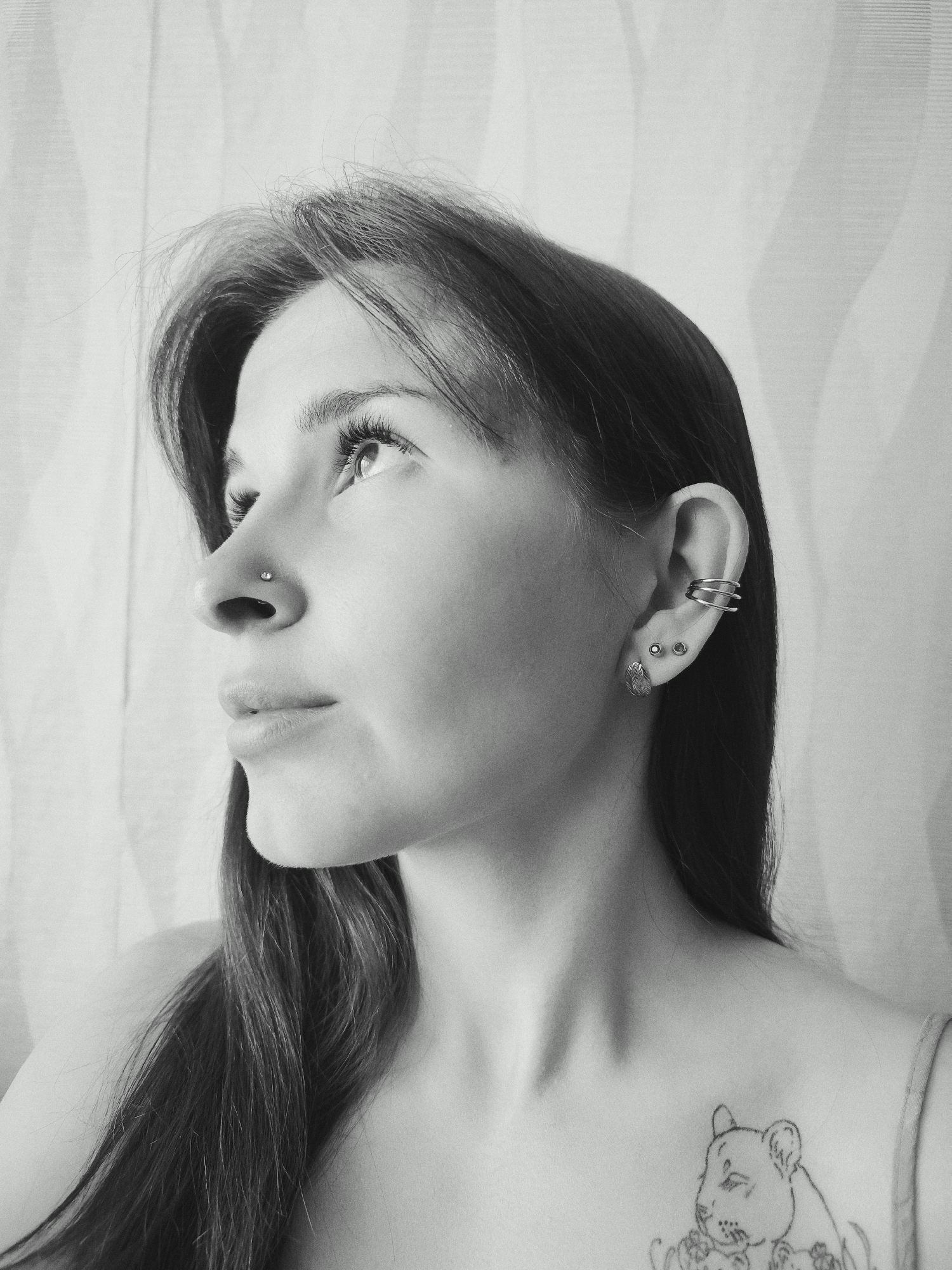 A black and white, medium shot image shows a young woman from the chest up. She has long, dark hair and is wearing a nose ring, ear cuff, and earrings. There is a tattoo of a lion cub on her upper left chest. Her head is turned to the right and she is looking upward. The background is a vertically striped pattern. There is no indication of the city of Фоки, Russia in the image.