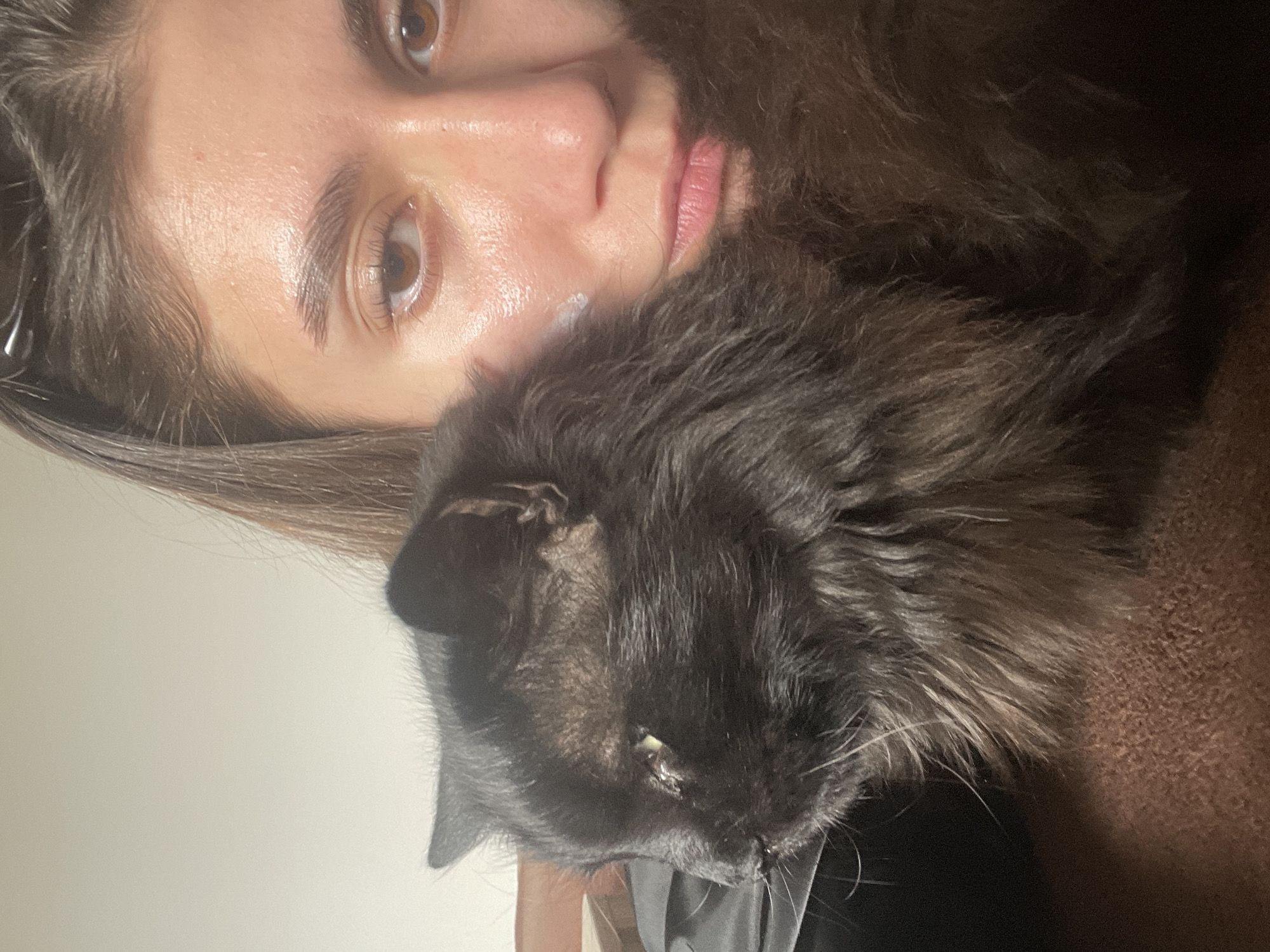 A close-up portrait, captured with a 90-degree counter-clockwise rotation, features a person and a black cat in an intimate setting. The human subject's face occupies the left side of the frame. She has warm brown eyes that gaze slightly past the viewer, dark, well-defined eyebrows, and long eyelashes. Her skin appears clear and slightly dewy, with a small visible dab of white cream or product on her cheek/chin area. Her lips are naturally pink and slightly parted. Dark brown hair, possibly tied back, frames her face and head.
Resting closely beside the person's face, primarily on the right side of the frame, is a fluffy black cat. The cat's head is tilted, with its left eye visible, showing a small amount of light-colored discharge near the inner corner. Its fur is dense and dark, with some lighter greyish strands visible, and prominent white whiskers extend from its muzzle. The cat appears calm, possibly resting or looking downwards.
The background on the far left is a plain, light-colored wall. Towards the right and beneath the cat, a textured brown fabric, possibly a blanket or a piece of furniture, is visible. A dark fabric, possibly clothing, can be seen underneath the cat's head. The scene suggests a peaceful, indoor environment, likely a home in Sofia, Bulgaria. The soft, ambient lighting suggests an evening or indoor setting without direct sunlight. There is no visible text in the image.