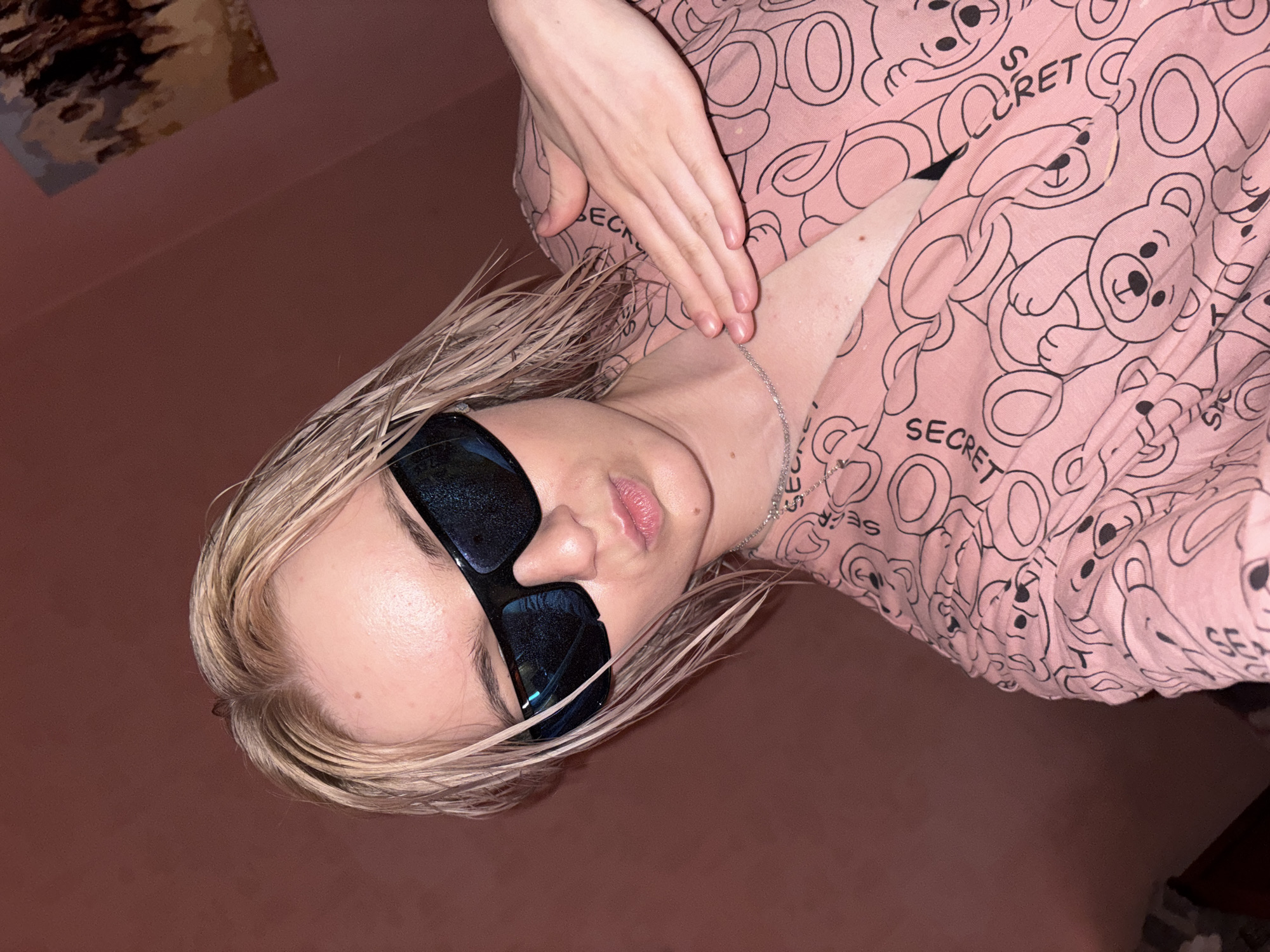 A person with blonde hair and dark, reflective sunglasses is captured in an angled indoor portrait, possibly a selfie. They are wearing a pink top adorned with a pattern of teddy bears and the repeated word "SECRET." A silver chain necklace is visible around their neck, and one hand rests gently on their chest or collarbone. Their lips are slightly parted, but their expression is largely obscured by the sunglasses. The background consists of a solid reddish-brown wall, with a faint, blurred artwork visible in the upper left corner. The lighting appears dim and artificial, suggesting an indoor setting without direct natural light. The location is specified as unknown, Russia.