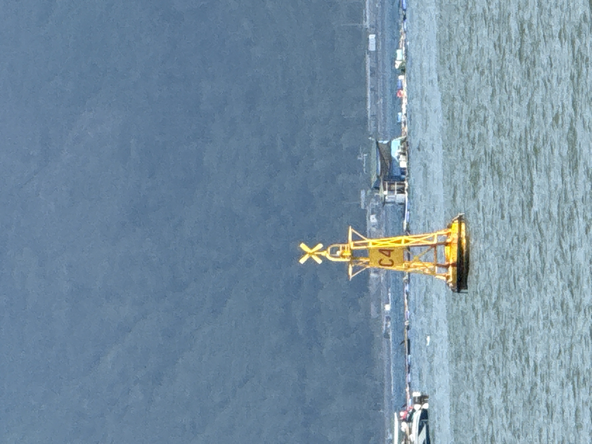 The image shows a yellow buoy in the water, with the marking "C4" on it. The buoy has a propeller-like structure at the top. In the background, there is a blue mountain range, and a body of water with several floating structures and boats. The weather appears to be clear. The scene is likely a bay or harbor area, possibly in Nam Nha Trang Ward, Vietnam, given the presence of aquaculture or fishing structures and the distinct mountainous backdrop.