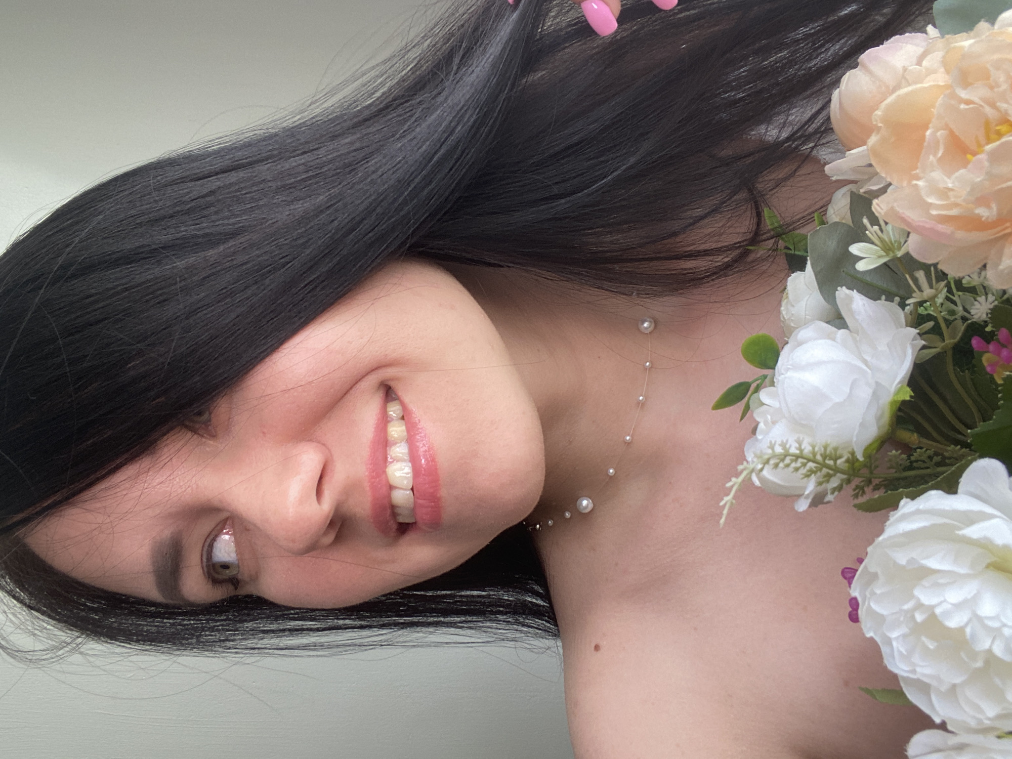 A young woman with dark hair and fair skin is shown from the chest up, smiling and looking to her left. She is wearing a sheer necklace with small pearls and holding a bouquet of white and peach flowers. Her nails are painted bright pink. The background is a plain, light-colored wall. There is no indication of the image being taken in Chaikovsky, Russia.