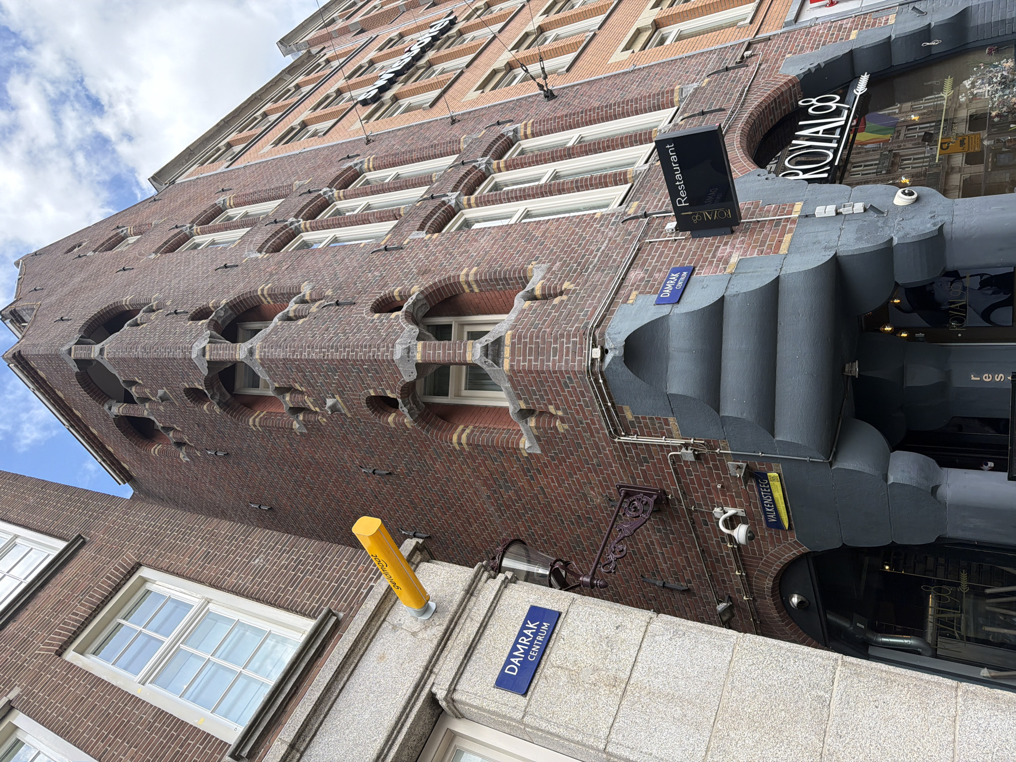 This image captures a street view in Amsterdam, Netherlands, focusing on the architecture of several adjacent buildings. The primary building in the center features a distinct brick facade with intricate, decorative stone elements framing arched windows. To the left, a building with a lighter colored, granite-like facade is visible, bearing a blue street sign that reads "DAMRAK CENTRUM". A bright yellow, cylindrical object, possibly a sign or part of a building's exterior feature, extends horizontally from this building. Below this, a traditional street lamp with an ornate bracket is mounted on the brick building.
Further to the right, another brick building with a more uniform facade is present, with multiple windows. A black sign with white lettering is mounted on this building, indicating "Restaurant". Below this sign, a larger neon sign reads "ROYAL 98" with an arrow graphic. The entrance below this sign shows reflections of other buildings, including a rainbow flag, suggesting it is a business, likely the restaurant mentioned. The sky above is partly cloudy. Several security cameras are visible mounted on the buildings, along with electrical wiring. The overall scene depicts a busy urban street environment with a mix of architectural styles.