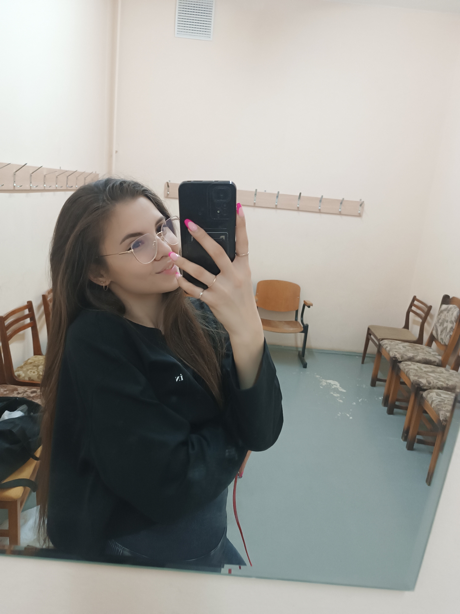 A woman is taking a selfie in a mirror inside a room. She has long brown hair, wears glasses, a black sweatshirt, and pink nail polish. She holds a black smartphone in her right hand, which is positioned towards the mirror. The room has light peach-colored walls with a row of hooks along one wall. Several wooden chairs are scattered around, including a tan wooden chair with black armrests. The floor is a light blue or gray color. A ventilation grille is visible on the wall near the ceiling.