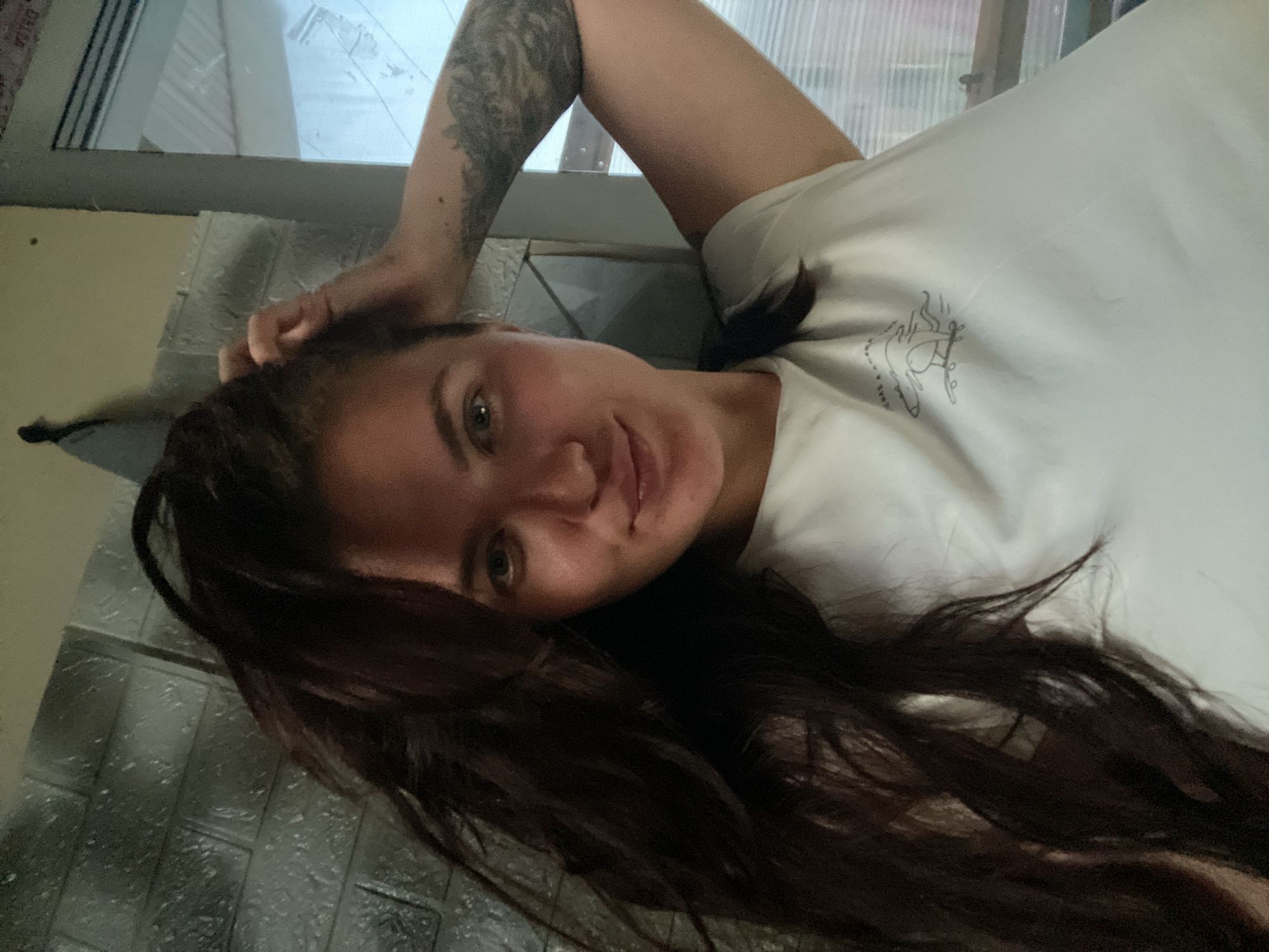 A woman is reclining indoors. She has fair skin, dark brown hair, and light-colored eyes. She is wearing a white t-shirt and has a tattoo on her left arm. She is looking at the camera, with her head tilted. The background appears to be a wall and a window. The location is Мосметаллоконструкция Онт, Russia.