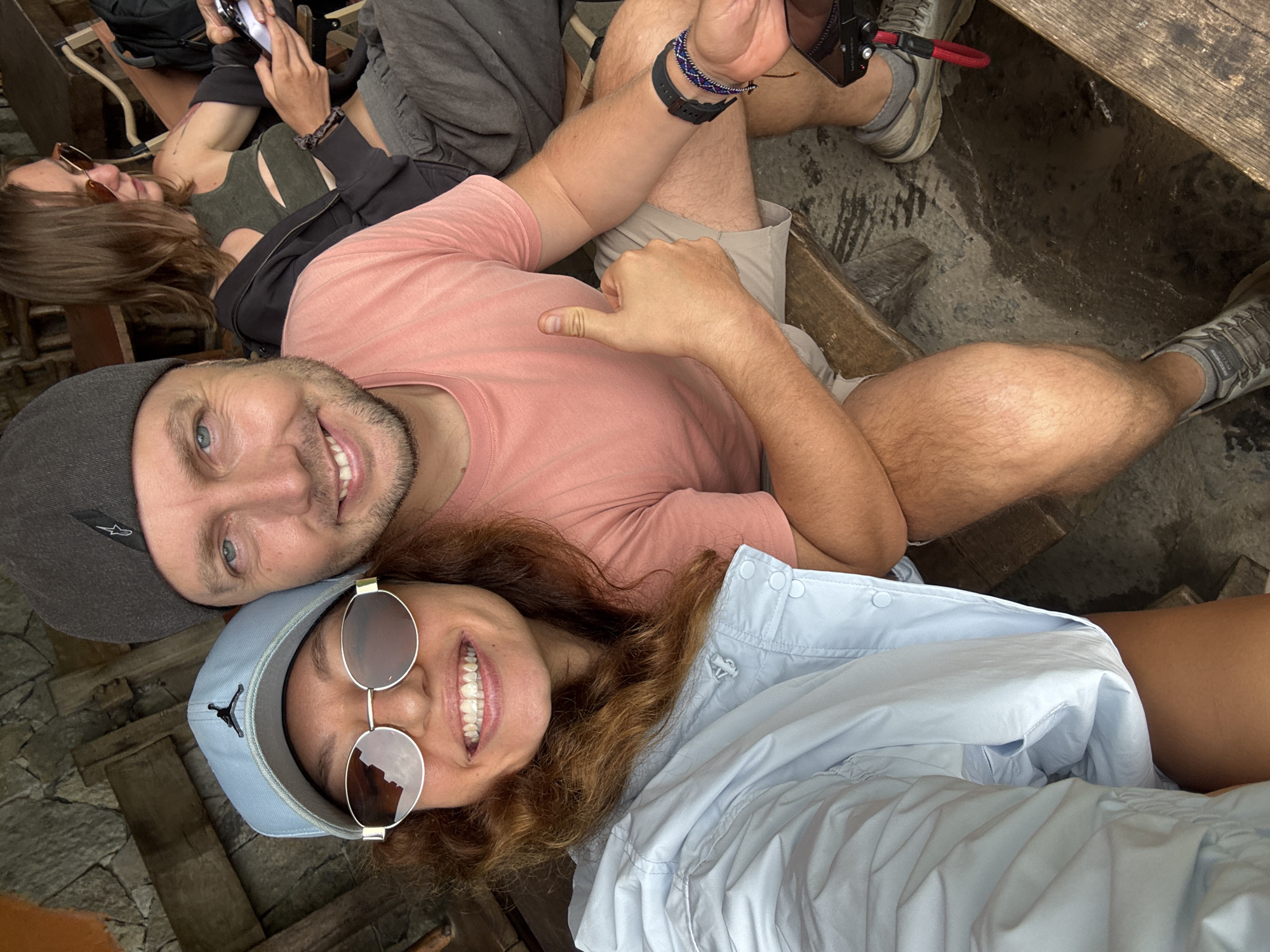 A group of people are resting in an outdoor setting. A man wears a gray cap and a pink shirt, while a woman sports a light blue cap and circular sunglasses. The woman wears a light blue shirt and is smiling. Another person is seen partially in the background. The setting appears to be a rest stop along stone walkways. Location: possibly in or around Yên Minh Commune, Vietnam.