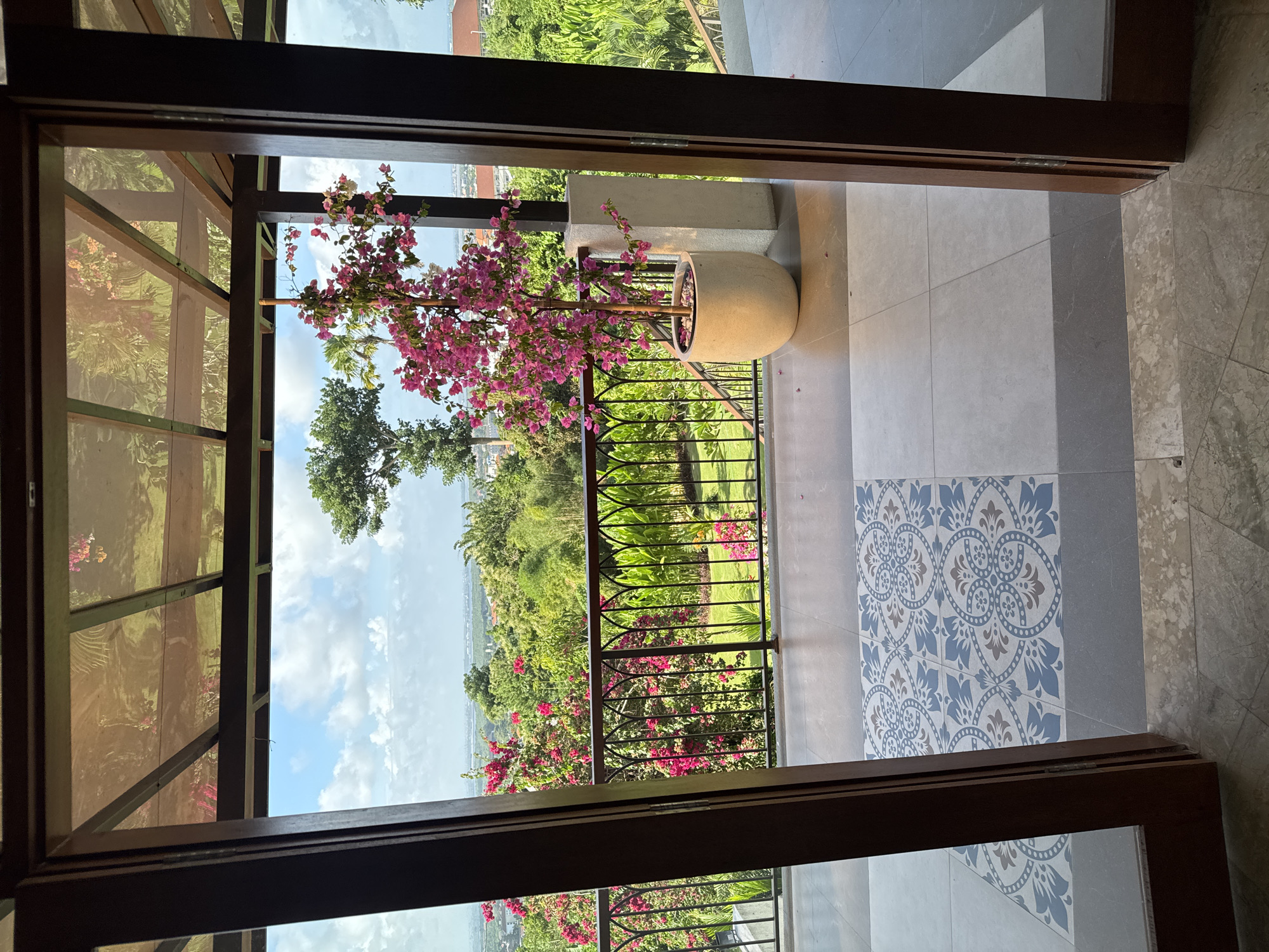 The image captures a view from inside a building or room, looking out through an open doorway or large opening. The location is Kuta Selatan, Indonesia.
The opening frames a scenic vista featuring a lush, green landscape, a distant body of water or ocean, and a partially visible sky. The foreground features a balcony or terrace area with wrought iron railings. On the balcony, there is a large potted plant with vibrant pink flowers, possibly bougainvillea, growing in a cream-colored pot.
The interior space appears to have tiled flooring, with a section of decorative blue and white patterned tiles near the doorway. The doorway itself is framed by dark wood, providing a contrasting border to the bright, open view beyond. The building or room seems to have a transparent or screened enclosure. The overall impression is one of a tropical, serene setting.