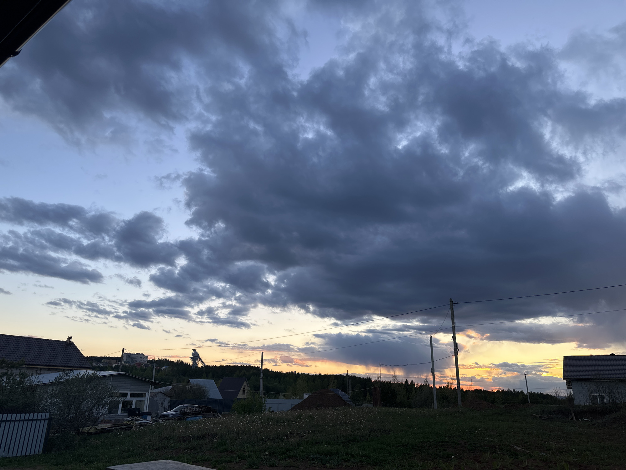 The image depicts a dusk or dawn scene in what appears to be a rural or suburban area of Прикамский, Russia. A large formation of dark, heavy clouds dominates the sky, tinged with a faint orange glow on the horizon indicating a setting or rising sun. Below the clouds, there is a visible treeline and various houses, some with fences and visible building materials, suggesting recent construction or renovation. Utility poles and overhead power lines are visible, adding to the sense of being in a developed area. The foreground shows a grassy field or lawn. In the distance, what appears to be a construction site or industrial structure is visible, possibly a crane or an elevator. The overall atmosphere suggests either an approaching storm or the aftermath of one.