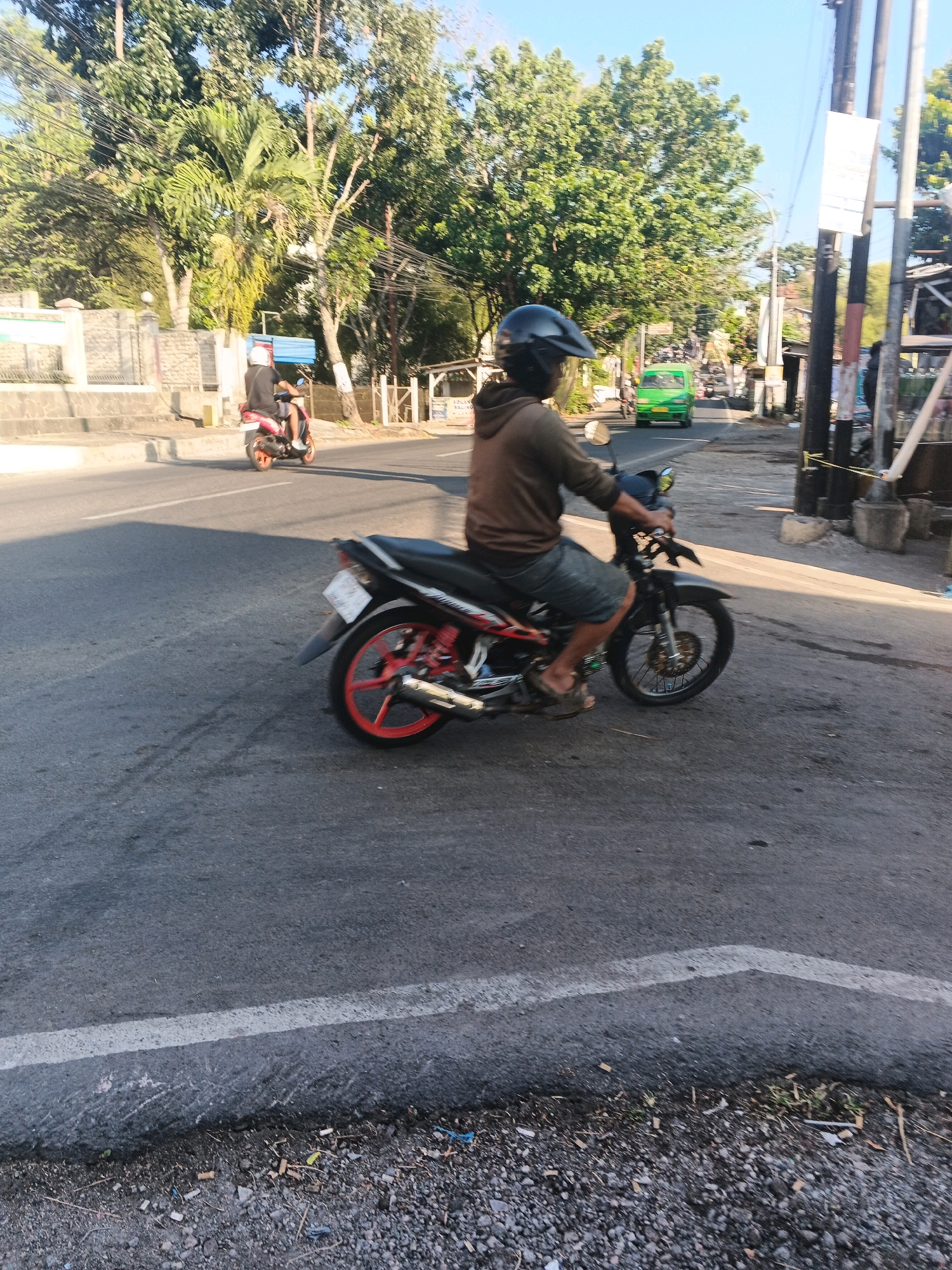 A medium shot captures a person on a red and black motorcycle riding on a paved road in Semarang, Indonesia. The rider, wearing a brown hooded sweatshirt, dark shorts, and a black helmet with a clear visor, is in the process of operating the vehicle. The motorcycle has a license plate that is not legible. In the background, another person on a red scooter is visible further down the road. A bright green van is also seen traveling in the opposite direction. The environment is a street with trees lining the sides and buildings in the distance. The road surface is asphalt with a white line marking the lane. The scene is illuminated by sunlight, casting shadows on the road. The edge of the road is bordered by gravel and vegetation.