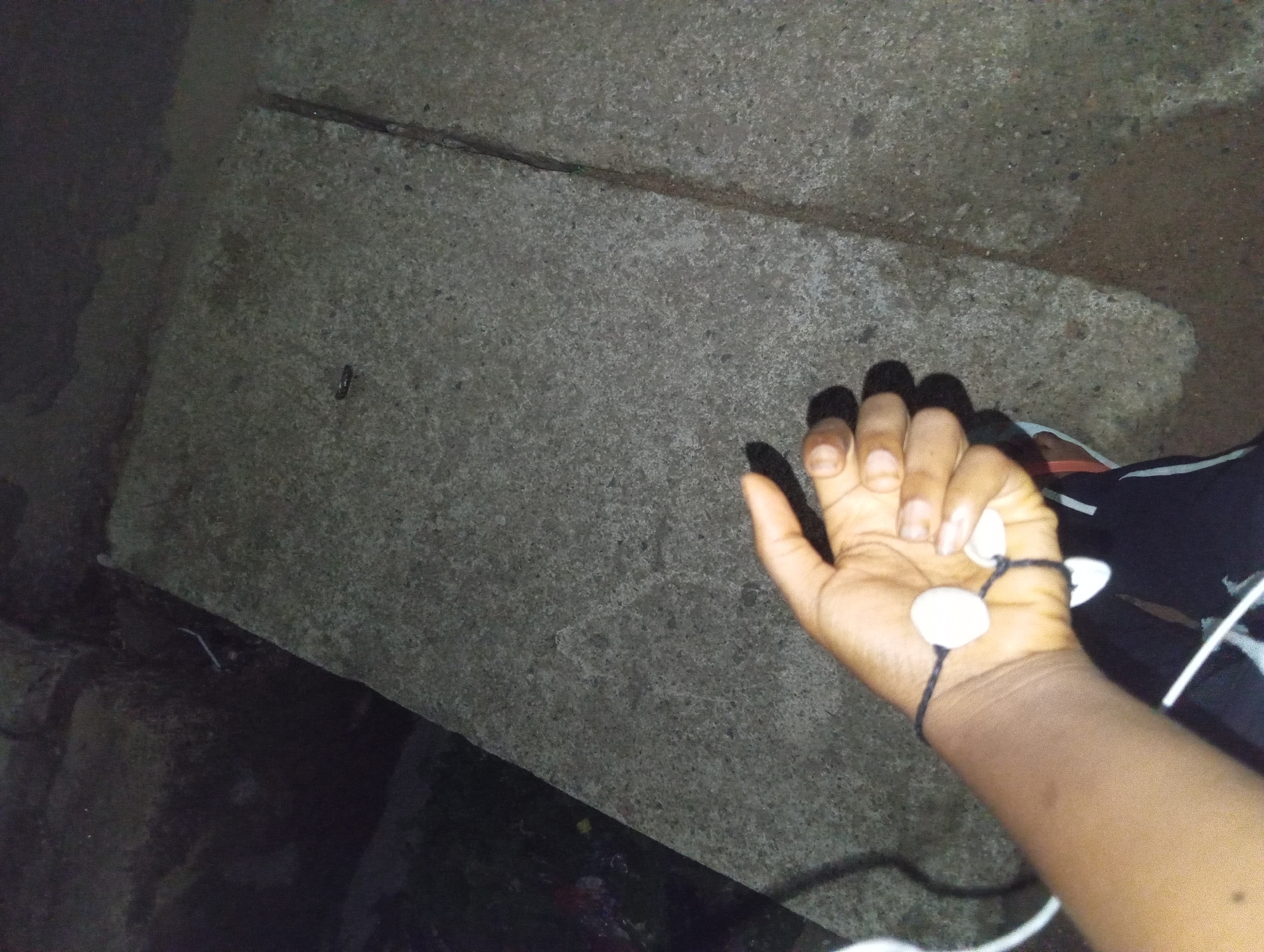 A person's left forearm and hand are visible in the lower left portion of the image. The arm is dark-skinned and is resting on a textured gray concrete surface that occupies the majority of the frame. A dark string bracelet with three white, teardrop-shaped beads is wrapped around the wrist. Two white earbud wires extend from the wrist area, disappearing out of frame to the left and bottom. The person is wearing black pants with white stripes down the side. The background consists of darker, rougher concrete or stone surfaces. The scene appears to be outdoors, possibly in an urban or park setting, given the concrete structures. The lighting is dim, suggesting it could be dusk, night, or an interior space with low light.