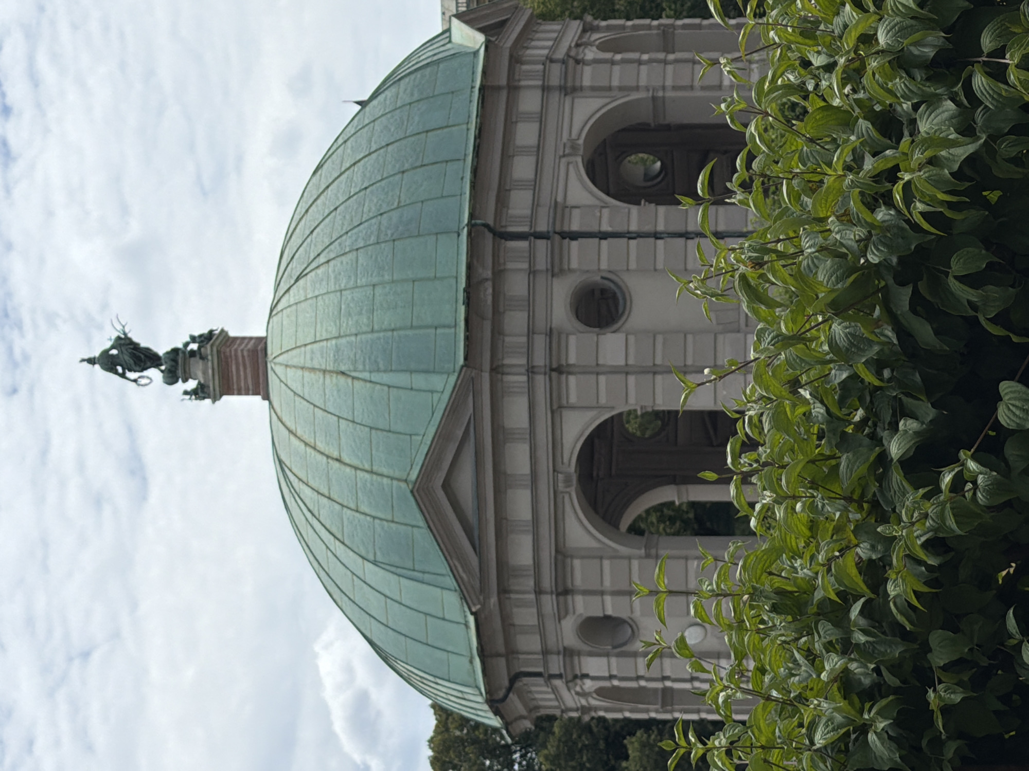 The image shows the Monopteros, a Greek-style temple located in the Englischer Garten in Munich, Germany. The temple features a copper-colored dome, a circular colonnade with arches, and a statue on top of the roof. In the foreground, there is a lush green hedge. The sky is cloudy. The Monopteros is a popular landmark in Munich, offering panoramic views of the city and the surrounding park.