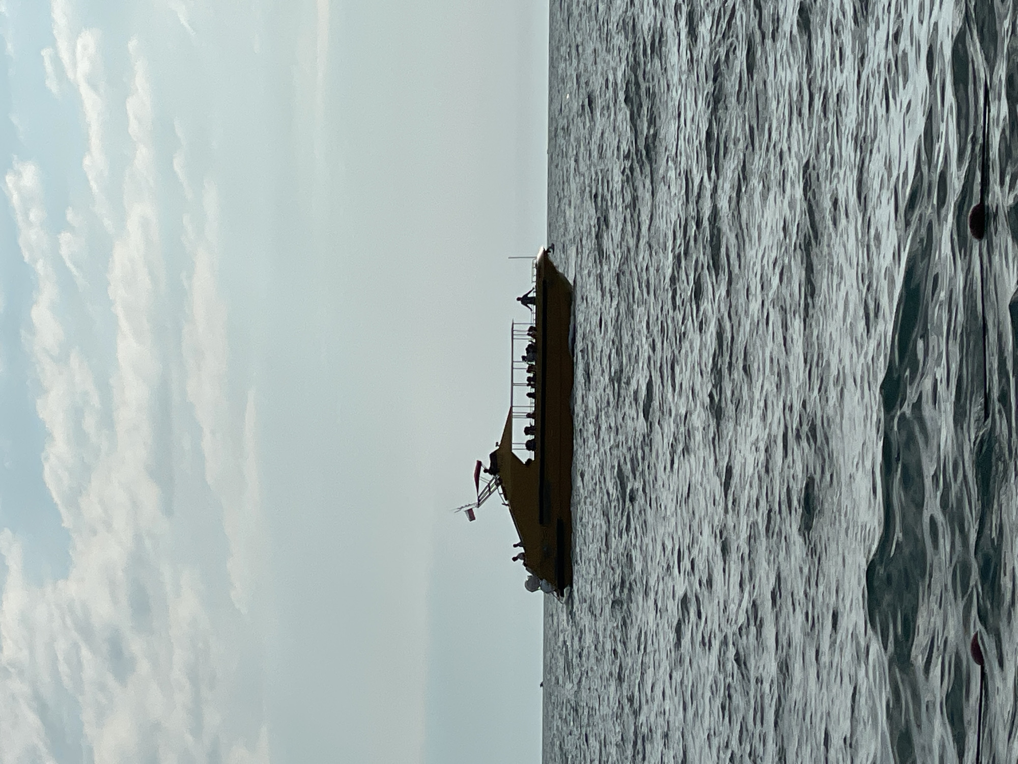 The image shows a bright, overcast day in Sochi, Russia. The composition is split vertically, with the left half depicting a pale blue sky filled with soft, white clouds, and the right half showing a choppy, gray sea. A yellow boat with several people on deck is positioned along the dividing line, oriented vertically in the frame. The boat appears to be a passenger vessel, with railings and a canopy visible. One person stands at the stern, looking out at the water. Two small flags, one red and white and another white, are affixed to a pole on the boat. The water is textured with numerous ripples and waves, suggesting a breezy condition. The light suggests it could be mid-morning or late afternoon. No visible text is present in the image. The scene evokes a sense of leisure or travel.
