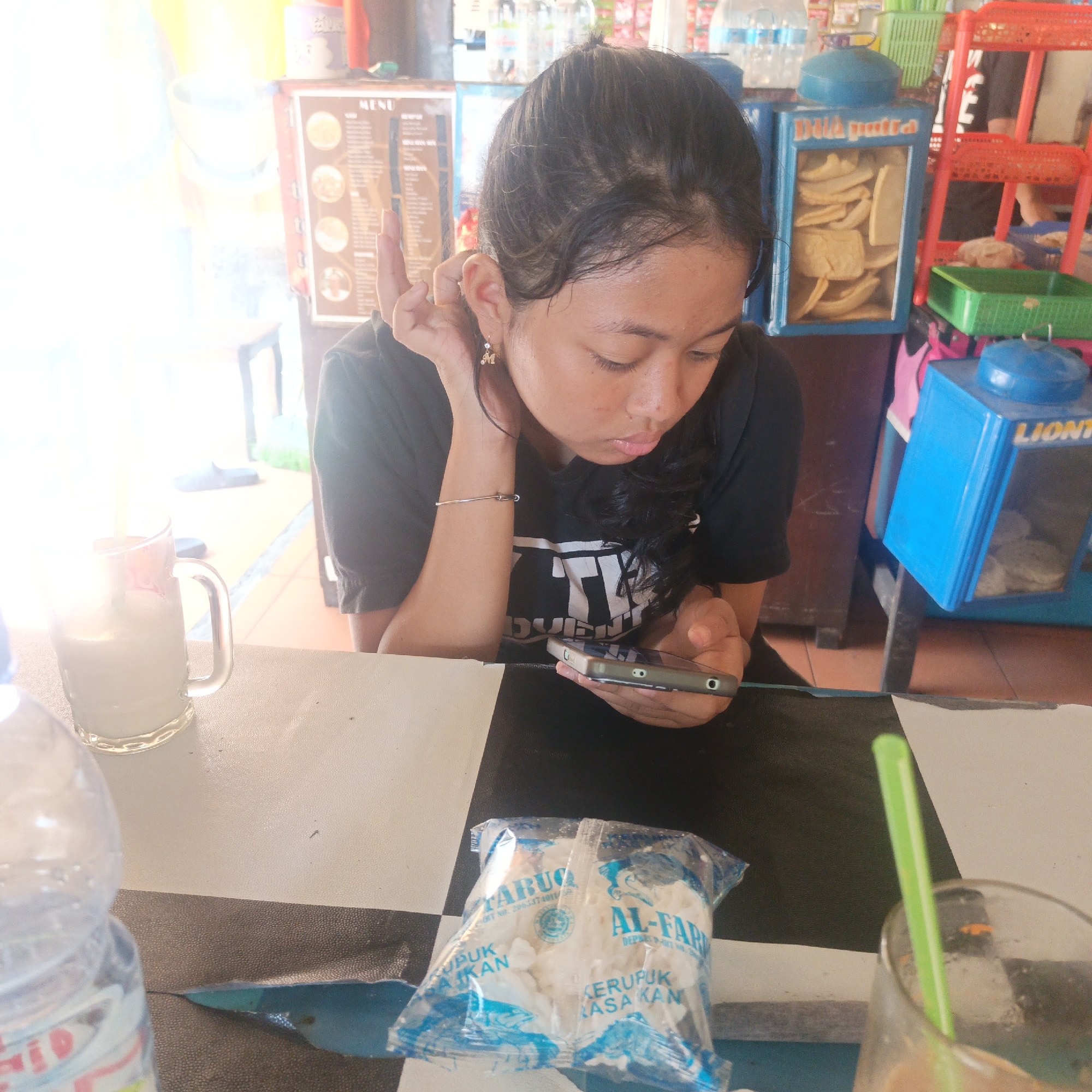 A young Indonesian woman with dark hair is seated at a table with a checkered pattern in a brightly lit, casual establishment. She is wearing a black t-shirt and appears to be looking down at her phone, which she holds in her right hand. Her left hand is resting on her ear. On the table in front of her is a glass of a light-colored beverage with a green straw, a bottle of water, and a clear plastic bag containing white crackers. In the background, there are shelves stocked with various items, including a blue cooler displaying snacks and a red rack. The overall environment suggests a small shop or food stall, possibly in Semarang, Indonesia, given the presence of local product packaging.