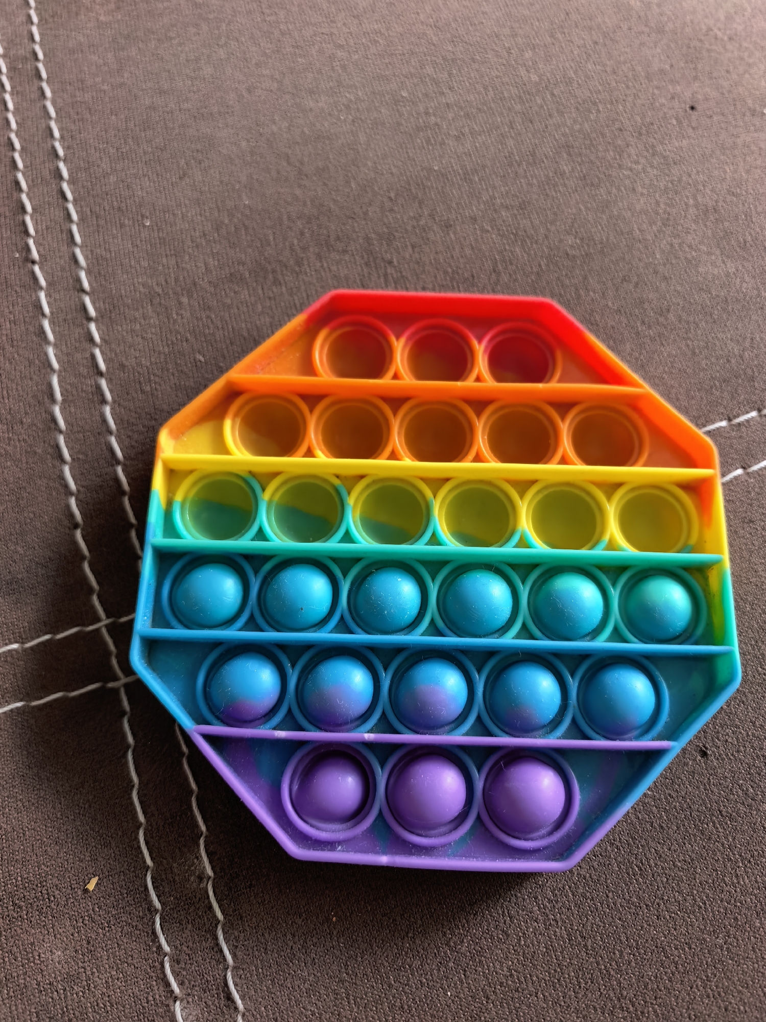 An overhead shot presents a multi-colored octagonal "Pop It" toy resting on a textured brown surface. The toy is comprised of rows of pliable bubbles in rainbow colors, including red, orange, yellow, green, blue, and purple. The background is a dark brown fabric material with a subtle texture and vertical lines of white stitching along the left side. There are no people or location-specific details available in the image.