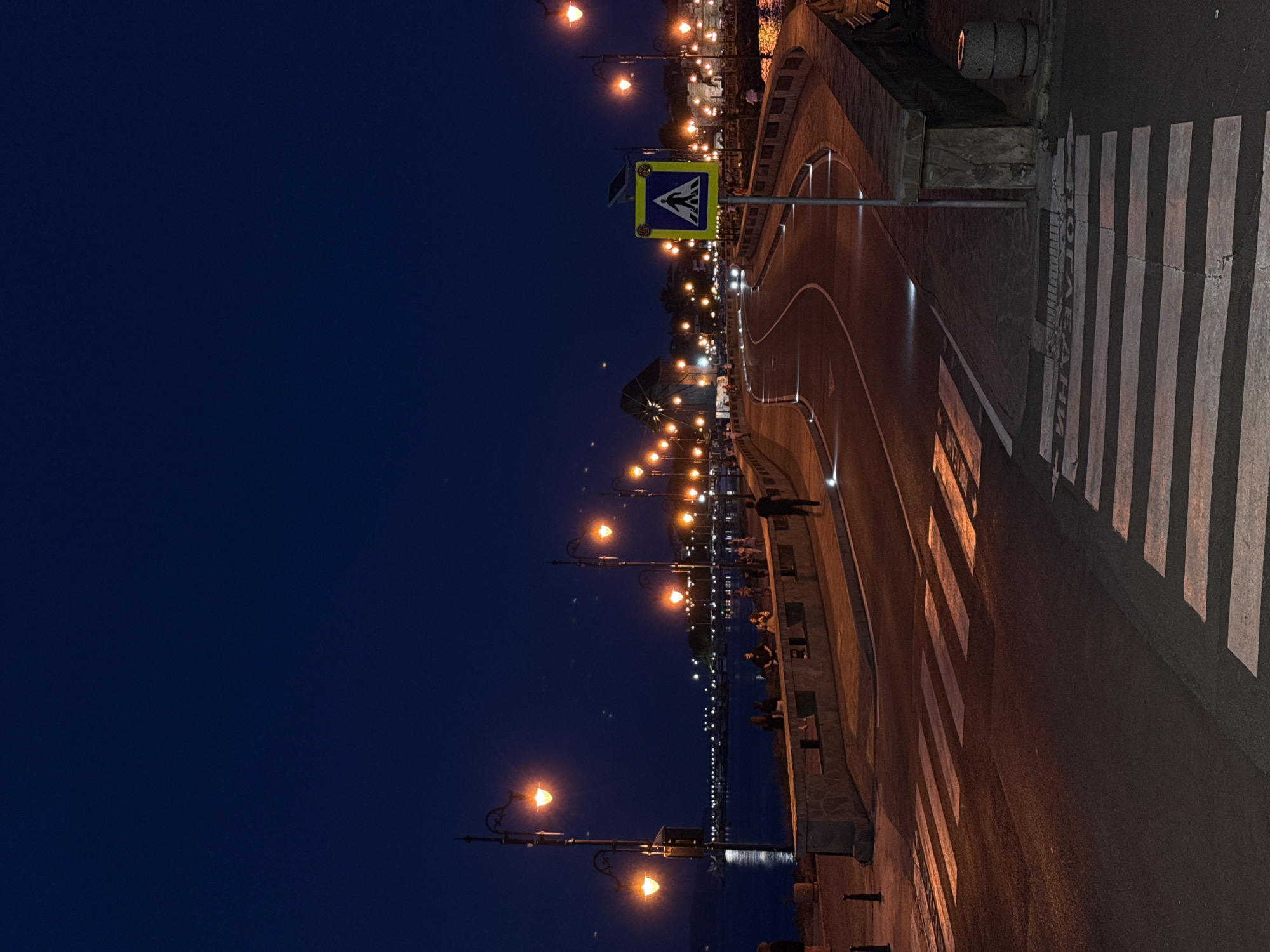 At dusk in Nesebar, Bulgaria, a pedestrian bridge with streetlights illuminates a coastal road. The road curves gently, with white stripes marking crosswalks. Several streetlights line the bridge, casting a warm glow. People are seen walking and sitting along the edge of the bridge, overlooking the sea. The water is dark, reflecting the lights of the distant town. A blue and yellow pedestrian crossing sign stands near the road. The sky is a deep blue, with a few faint stars visible.