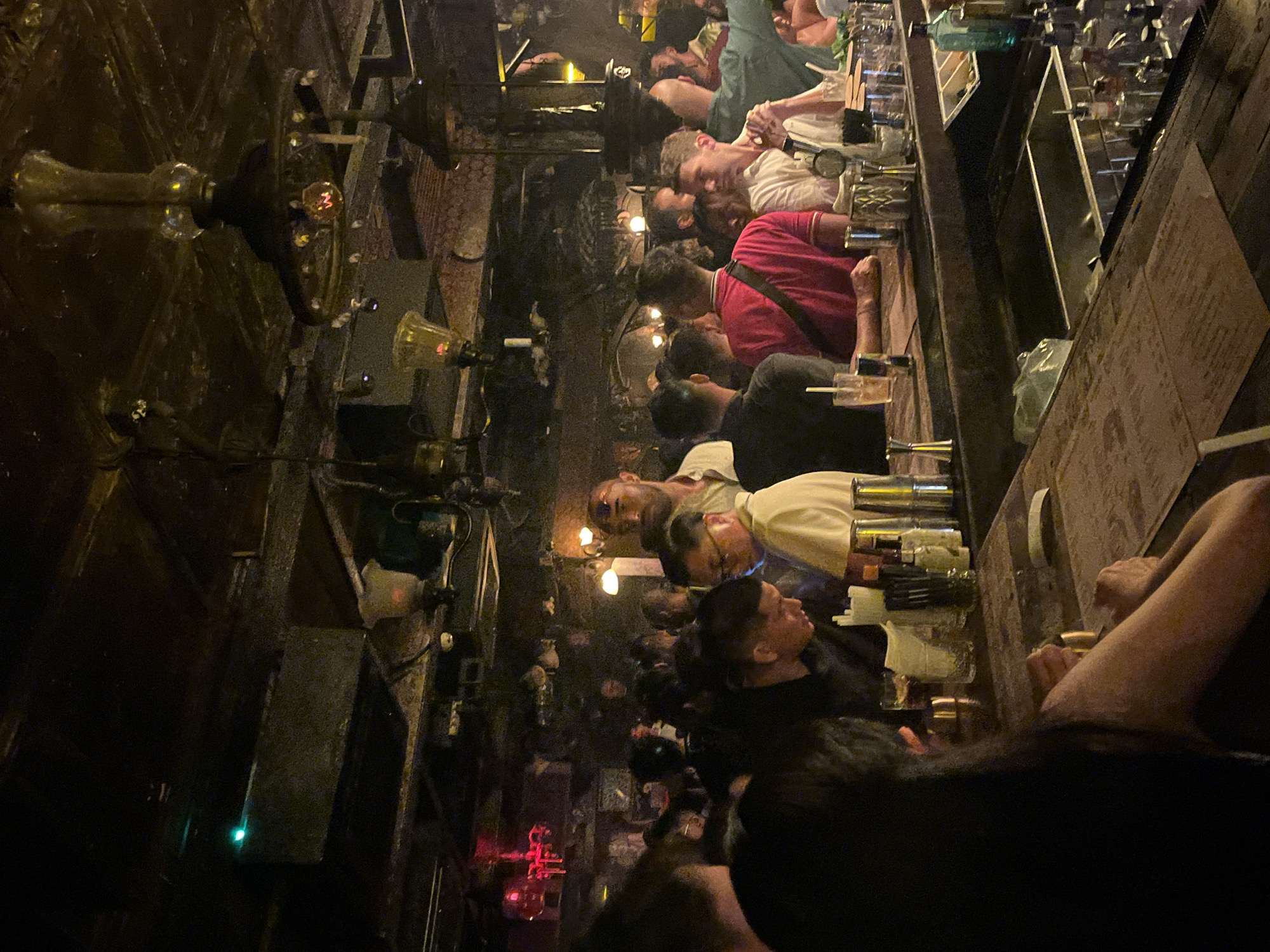 The image depicts a crowded indoor bar scene, likely in Kuta, Indonesia, characterized by dim lighting and a rustic, somewhat industrial aesthetic. The ceiling features exposed metal beams and ductwork, with various light fixtures including chandeliers and pendant lamps, some with ornate glass shades. In the background, a red light fixture is visible.
The bar area itself is busy with patrons and staff. Several people are standing around the bar, some appear to be ordering or receiving drinks, while others are engaged in conversation. The bar counter is made of wood and is lined with bottles, glasses, cocktail shakers, and other bar equipment. Some drinks with straws are visible on the counter.
In the foreground, a person with long dark hair is seated at the bar, with their back to the camera. Their arms are resting on the counter, and they appear to be interacting with something on the surface. The overall atmosphere suggests a lively nightlife setting.