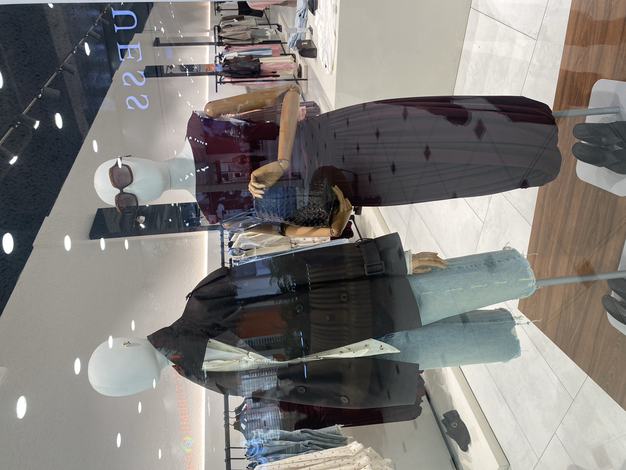The image shows a storefront window display, likely in a shopping mall or retail district in Yekaterinburg, Russia. Two mannequins are visible. The upper mannequin is wearing sunglasses, a dark red top, and a long, dark skirt. The lower mannequin is wearing a black leather jacket over a light-colored floral shirt with blue jeans. The display is situated in front of clothing racks with various garments. The window reflects the surrounding environment, including the ceiling, lighting fixtures, and the floor pattern of the interior space.