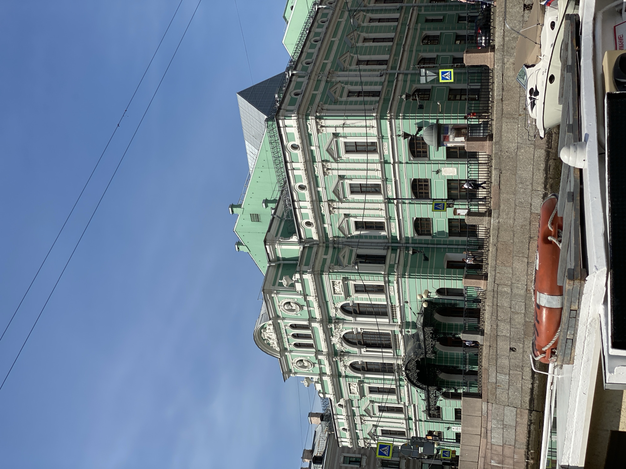 The image shows a bright, sunny day with a clear blue sky over a green building in Saint Petersburg, Russia. The building is ornate, with white trim and classical architectural elements. It appears to be a historic structure, possibly a theater or opera house, given its grand facade and decorative detailing. The building is situated along a canal or river, with a stone embankment. In the foreground, parts of a boat are visible, including an orange life preserver and a white motorboat docked alongside. People are seen walking along the embankment in front of the building. The scene captures a picturesque view of the city's architectural beauty and waterfront.