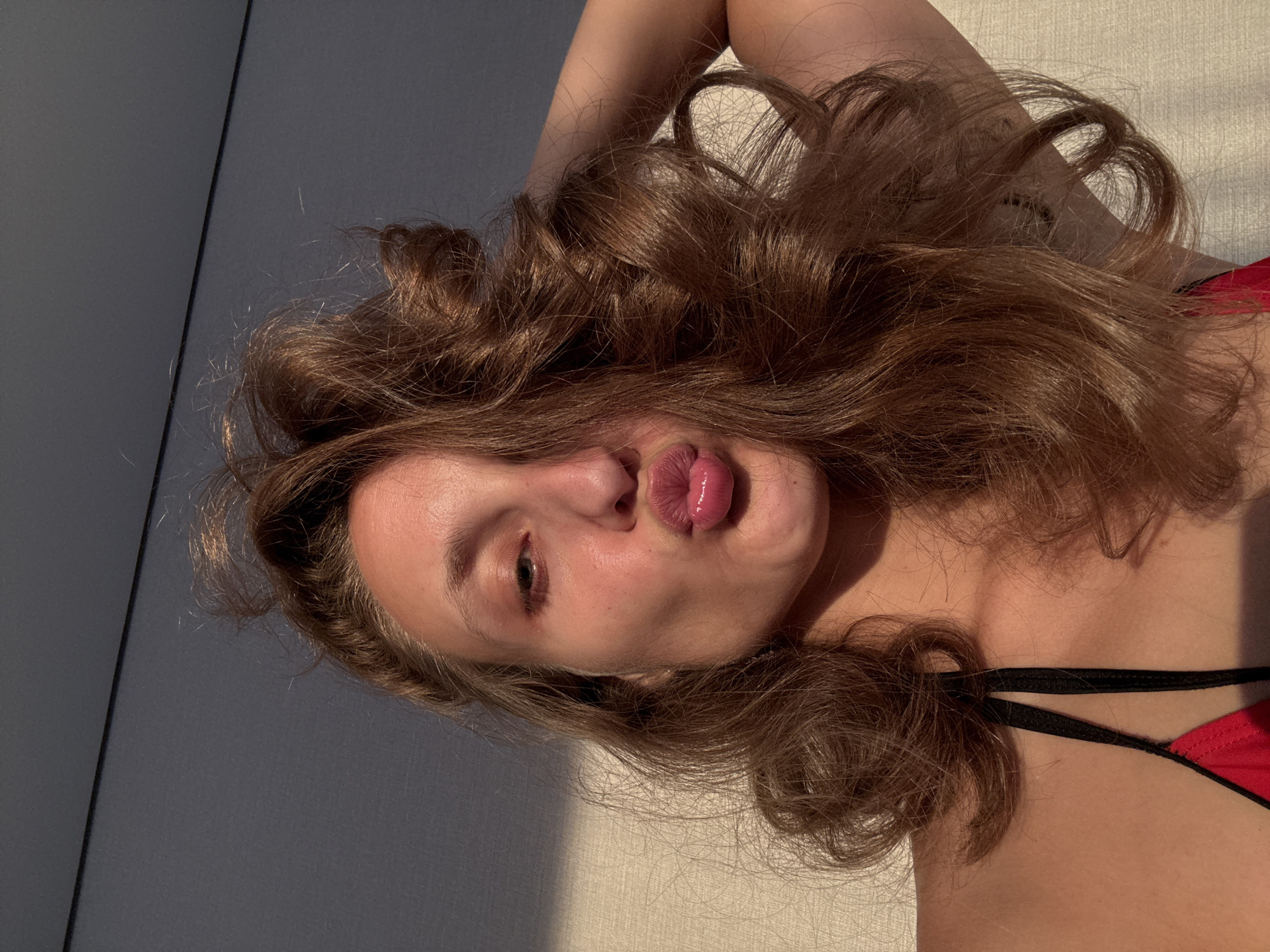 A young woman with long, wavy brown hair is posing for a selfie. She is wearing a red and black bikini top. Her face is turned towards the camera, and she is puckering her lips in a kissing gesture. The background appears to be a textured wall with shadows cast upon it, suggesting an indoor setting with natural light. The lighting suggests it is either early morning or late afternoon. There are no discernible elements in the image that can indicate its location within the city of Подшивалово, Russia.